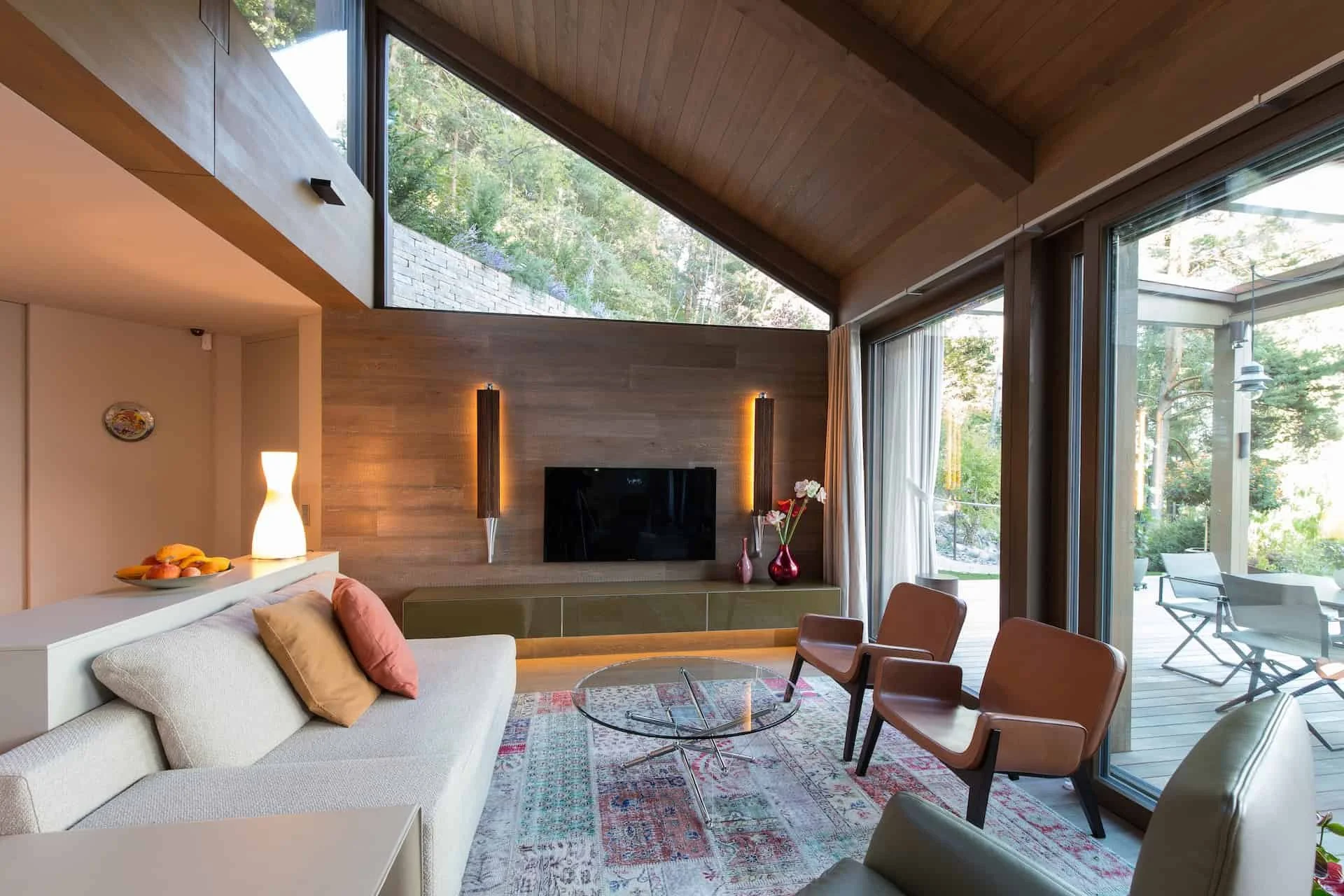Ein modernes Wohnzimmer mit großen Fensterfronten, Holzwänden und -decken, einer weißen Couch mit Kissen, einem Couchtisch, sechs Stühlen, einer Vase mit Blumen, einem Fernseher und einem Kamin. Draußen sind Bäume und eine Terrasse sichtbar.