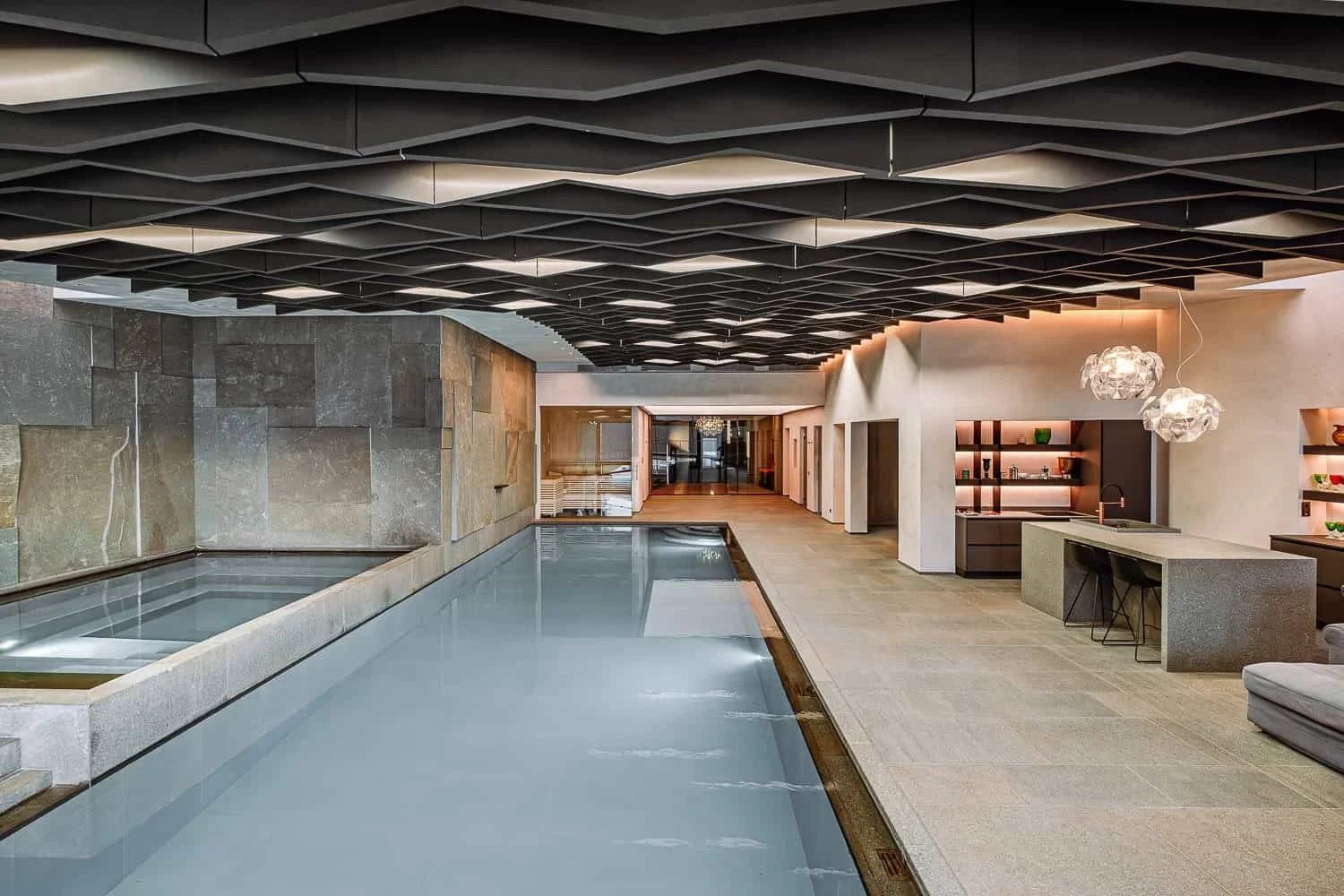 Innenansicht eines modernen Wellnessbereichs mit beheiztem Pool, Steinelementen an der Wand, Holzfußboden, luxuriösen Lampen und dekorativen Regalen.