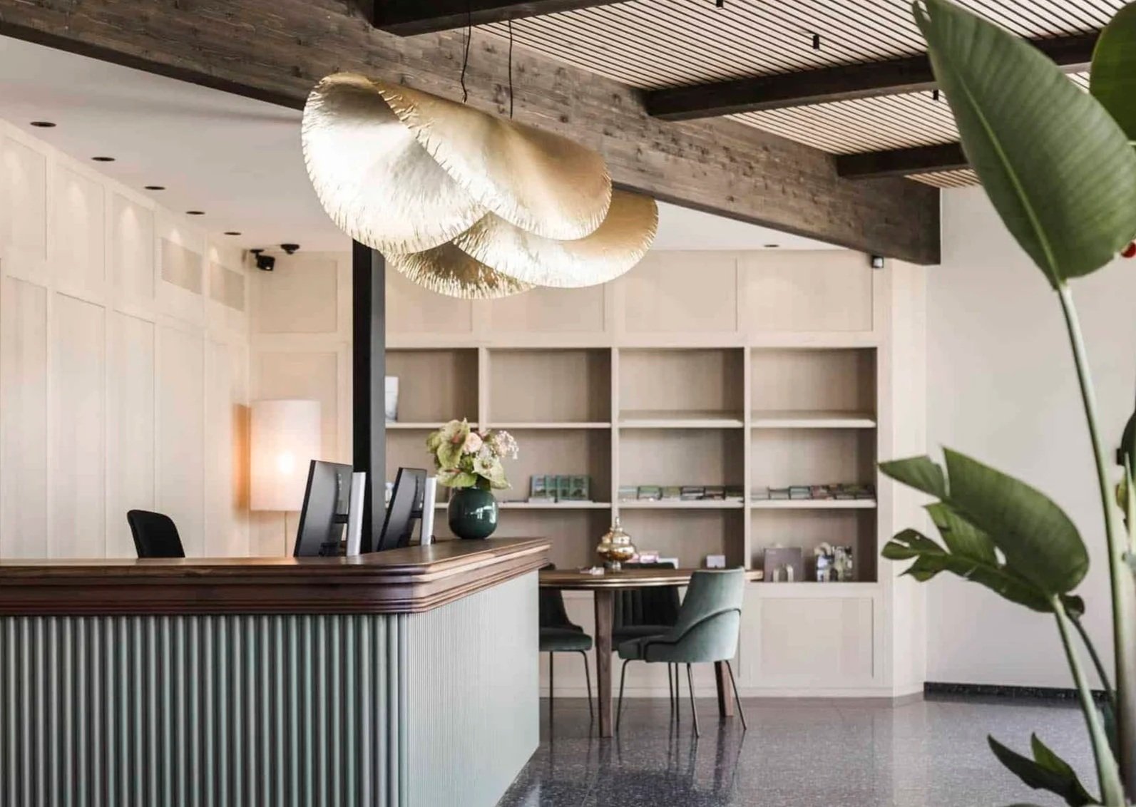 Ein stilvolles Hotel- oder Empfangsbereich mit Holz und cremefarbenen Wänden, einer kleinen Bar mit Stühlen, einer Blumenvase auf der Bar, einem Sofa, einem Bücherregal im Hintergrund und einer großen Zimmerpflanze im Vordergrund.