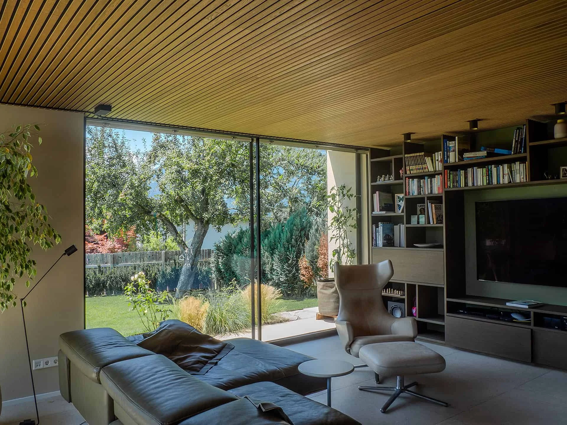 Geräumiges Wohnzimmer mit großen Fensterfronten, Blick auf einen Garten mit Bäumen, moderne Einrichtung mit Bücherregal und bequemen Sitzmöbeln, Holzdecke.