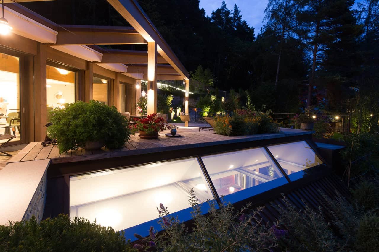Ein Abendbild einer modernen Terrasse mit vielen Pflanzen, großen Fenstern und einem beleuchteten Dachfenster, umgeben von Bäumen in der Dämmerung.