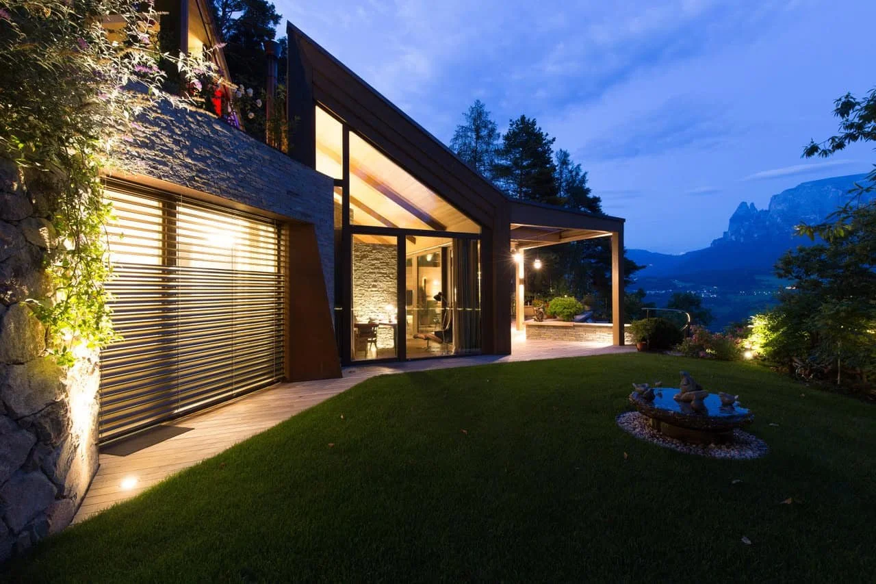 Modernes Haus mit großen Glasfenstern und Terrasse bei Abenddämmerung, umgeben von Garten mit Rasen und dekorativen Steinen, im Hintergrund Berge und Bäume.