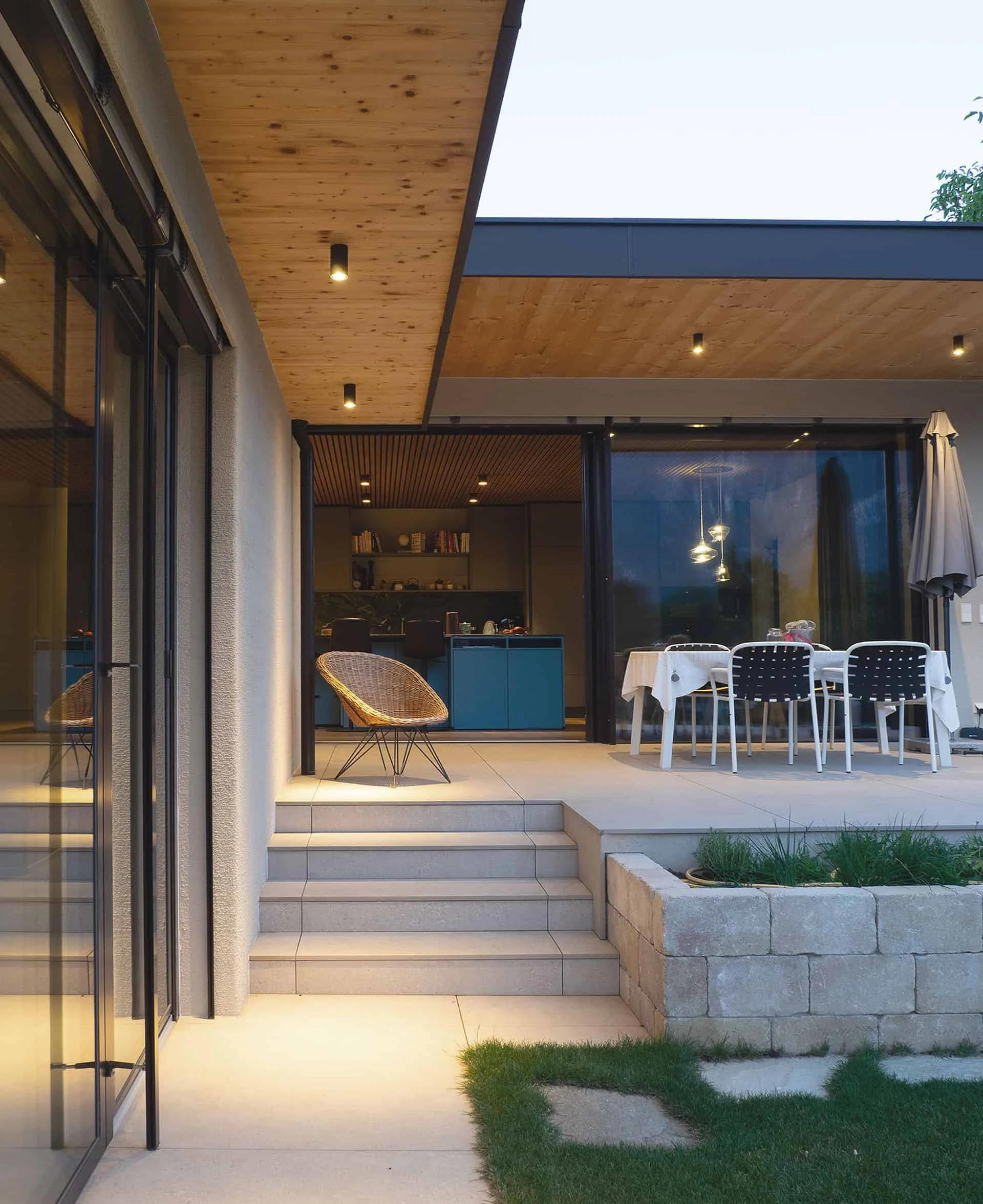 Moderner Außenbereich eines Hauses mit Terrasse, Stufen, Garten, large Fenster und Holzdach, inklusive Gartenmöbel und Beleuchtung.