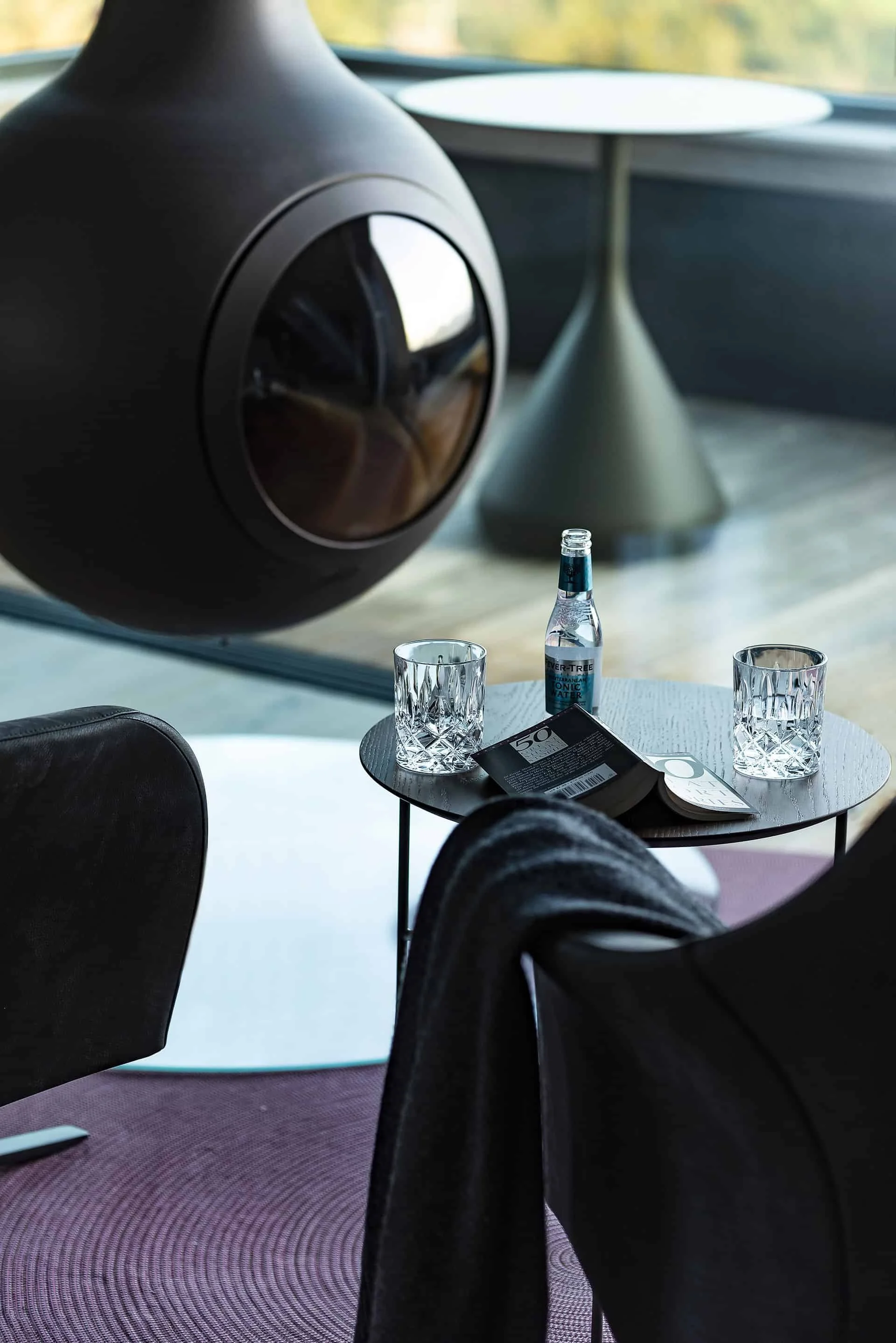 Innenaufnahme eines modernen Raumes mit schwarzer dekorativer Lampe, einem Tisch mit Wasserflasche, Gläsern und Zeitschriften, und schwarzen Sesseln auf einem karierten Teppich.