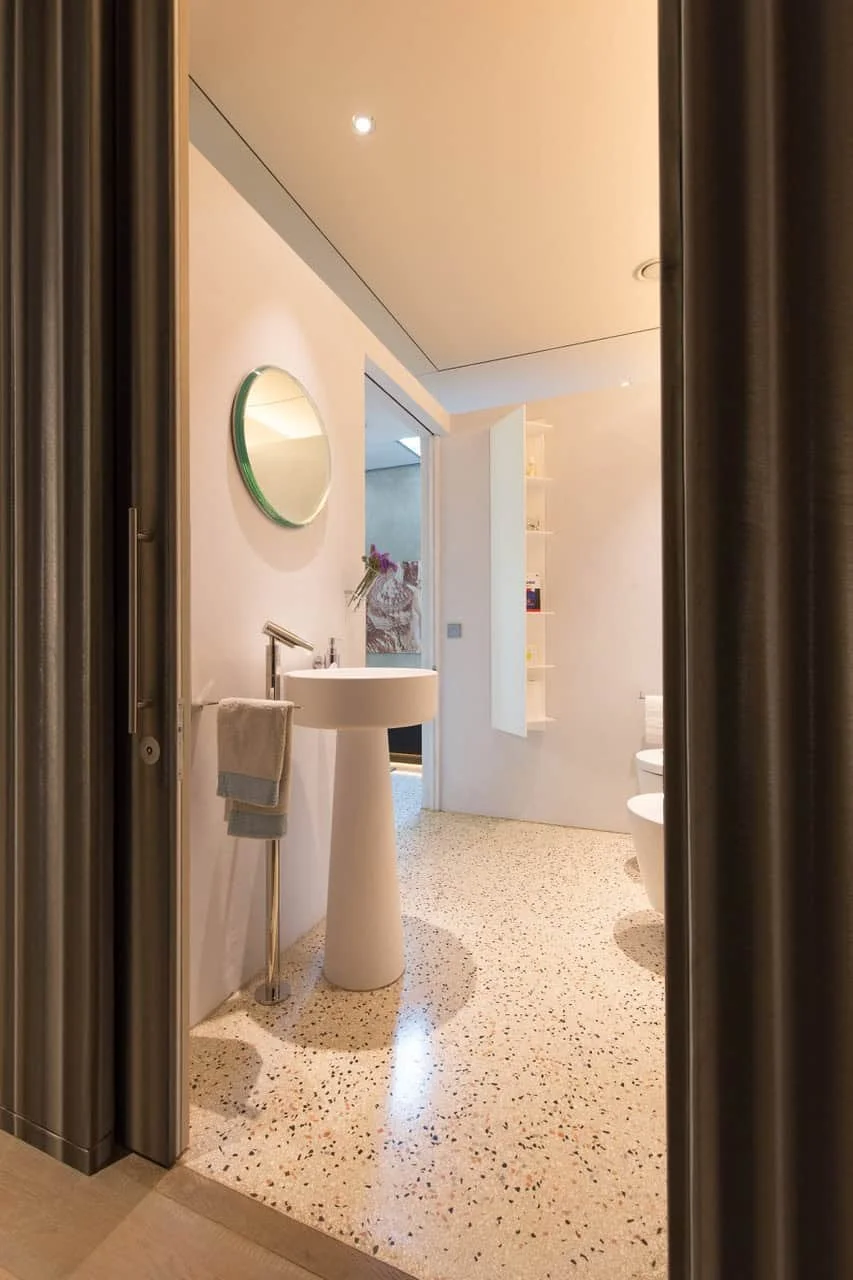 Schlichtes modernes Badezimmer mit rundem Spiegel, Waschbecken auf Säule, Handtuchstange, und einer weißen Toilette, sichtbar durch eine offene Tür.