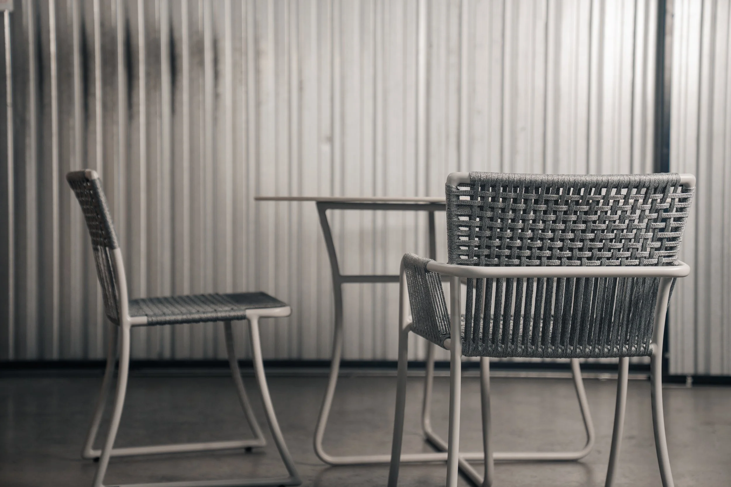 Three modern outdoor chairs with woven backs and seats around a glass table, set against a corrugated metal wall.