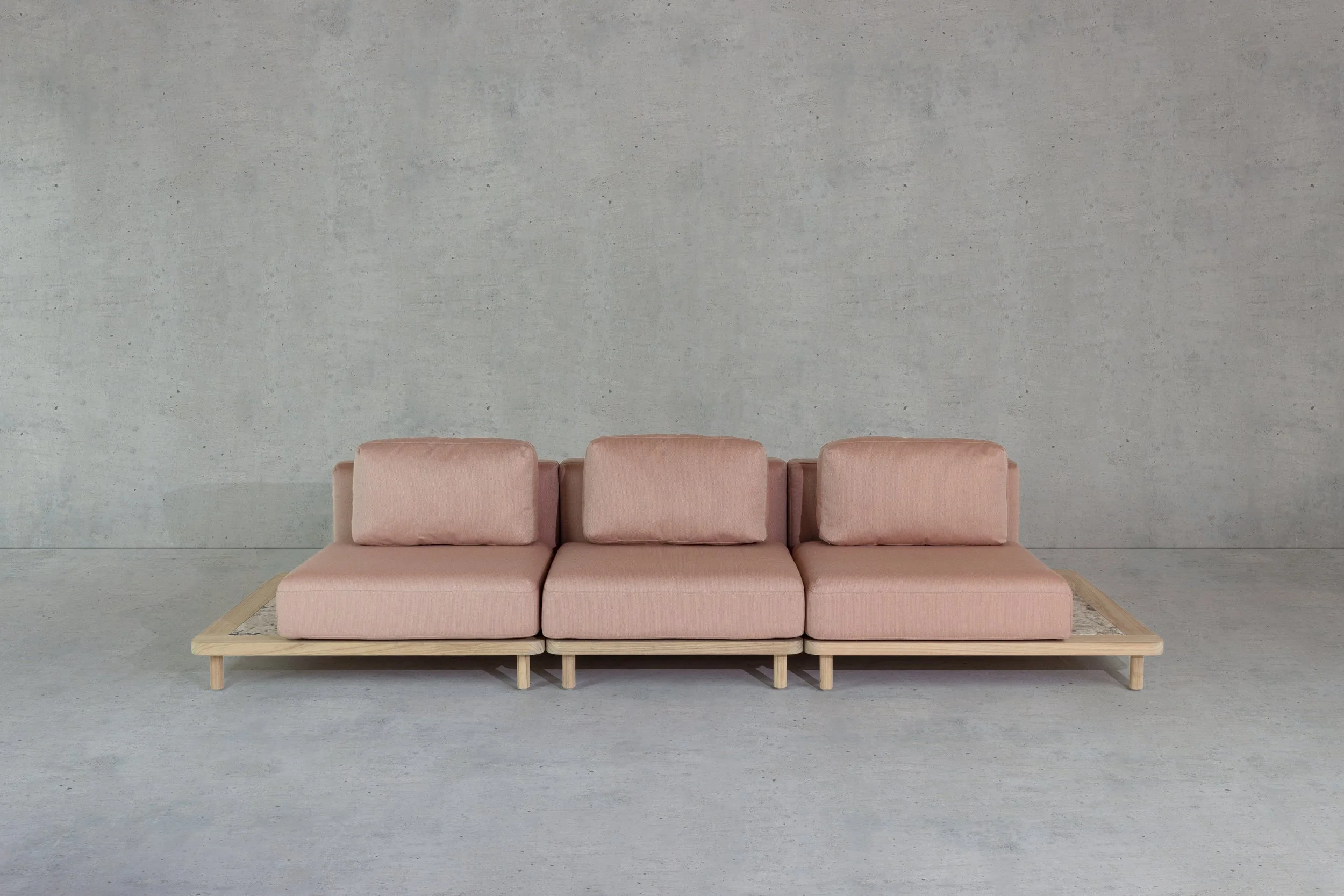 A modern, minimalist pink sofa with cushions, placed on a wooden frame, in front of a plain gray concrete wall.