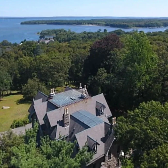 An aerial view of a large Victorian-style house with gray roofs, surrounded by green trees. In the background, there is a body of water and a forested landscape extending to the horizon.
