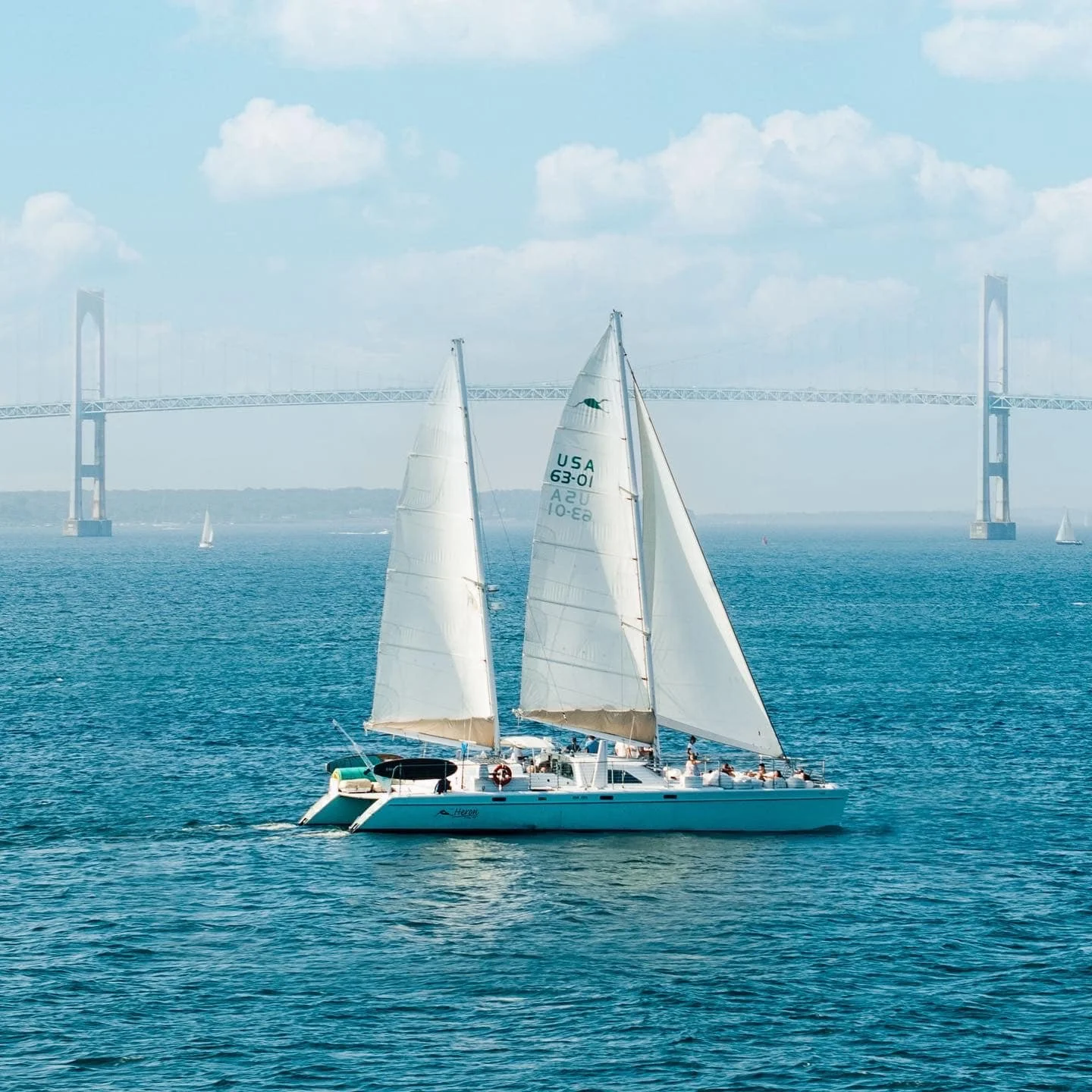A sailboat with white sails on the water, with a bridge in the background.