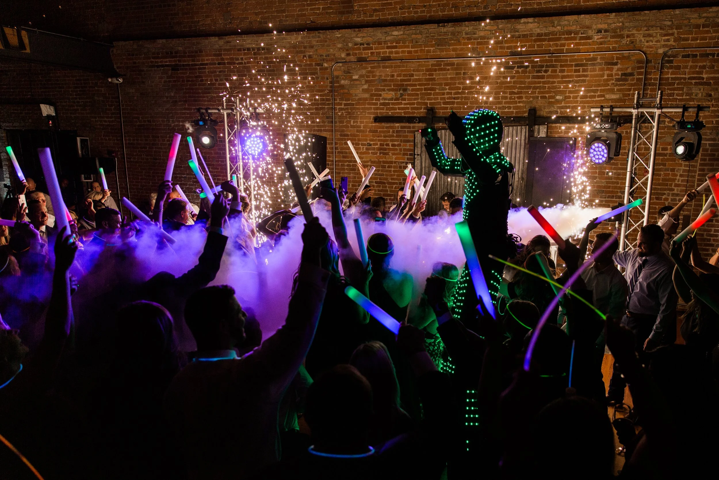 Crowded party with people holding glow sticks and a performer in an LED suit, surrounded by smoke and light effects.