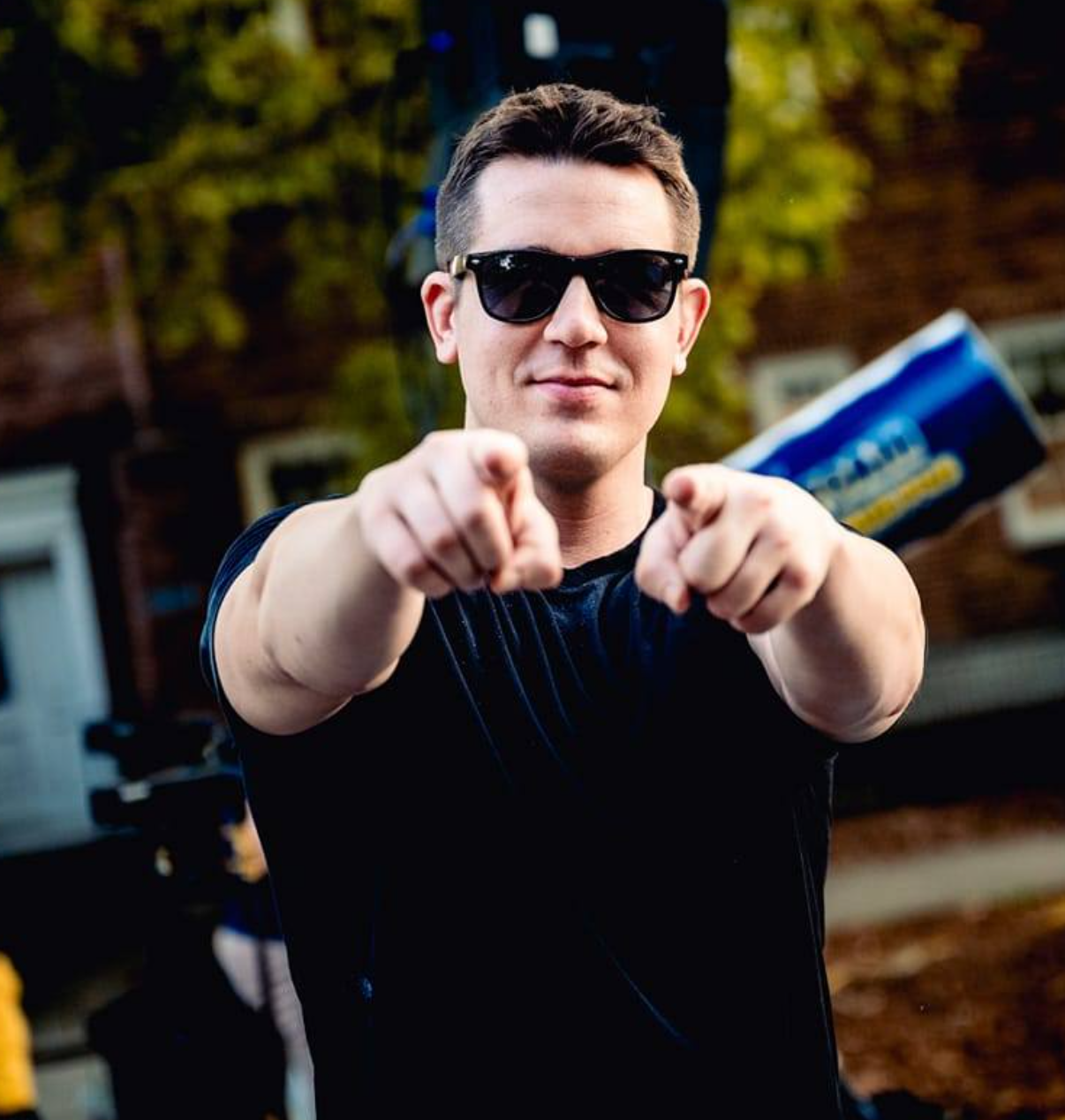 A person wearing sunglasses and a black shirt, smiling and pointing at the camera. They have a can under one arm. The background appears to be outdoors with trees and buildings.