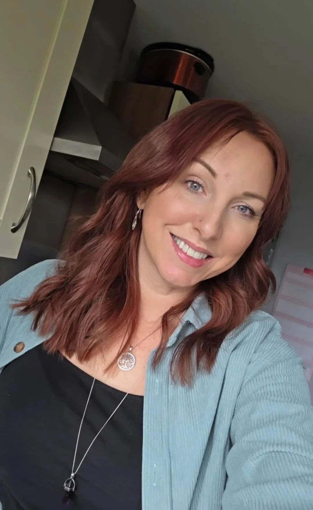 A life and mindset coach with red hair, smiling, wearing a black top and a light blue cardigan, taking a selfie in an indoor setting.