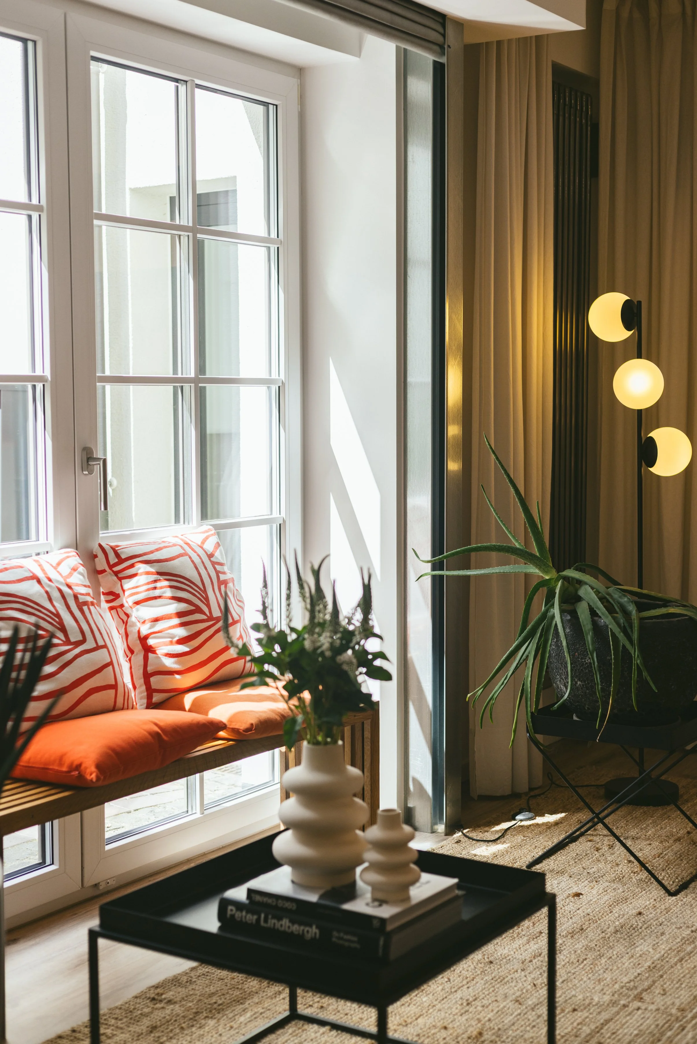Ein Wartebereich mit großen Fenstern, einer Bank mit orangefarbenen Kissen, einem schwarzen Beistelltisch mit Büchern und dekorativen Gegenständen, Pflanzen und einem Stehlampen-Design mit drei globusförmigen Lampen.