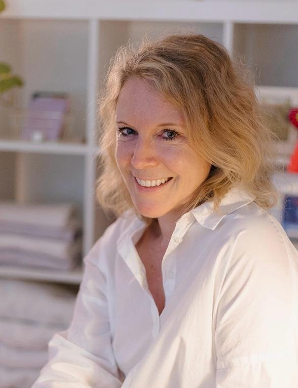 Lächelnde Frau mit lockigem, blondem Haar trägt ein weißes Hemd, im Hintergrund ein Bücherregal mit Akten und Dekorationen