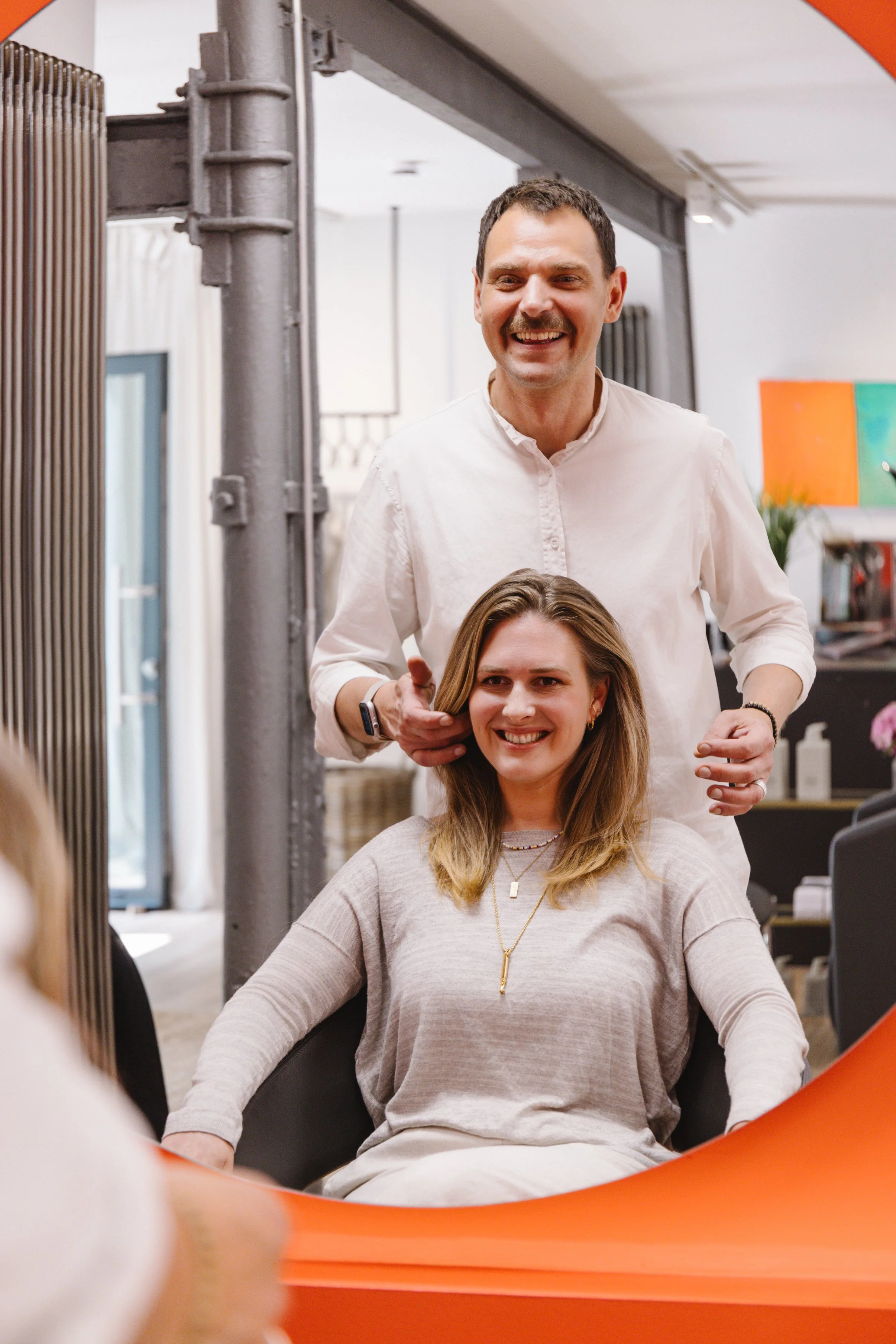 Ein Mann und eine Frau sitzen in einem Salon, während der Mann hinter der Frau steht und sie an den Haaren hält. Beide lachen und schauen freundlich.