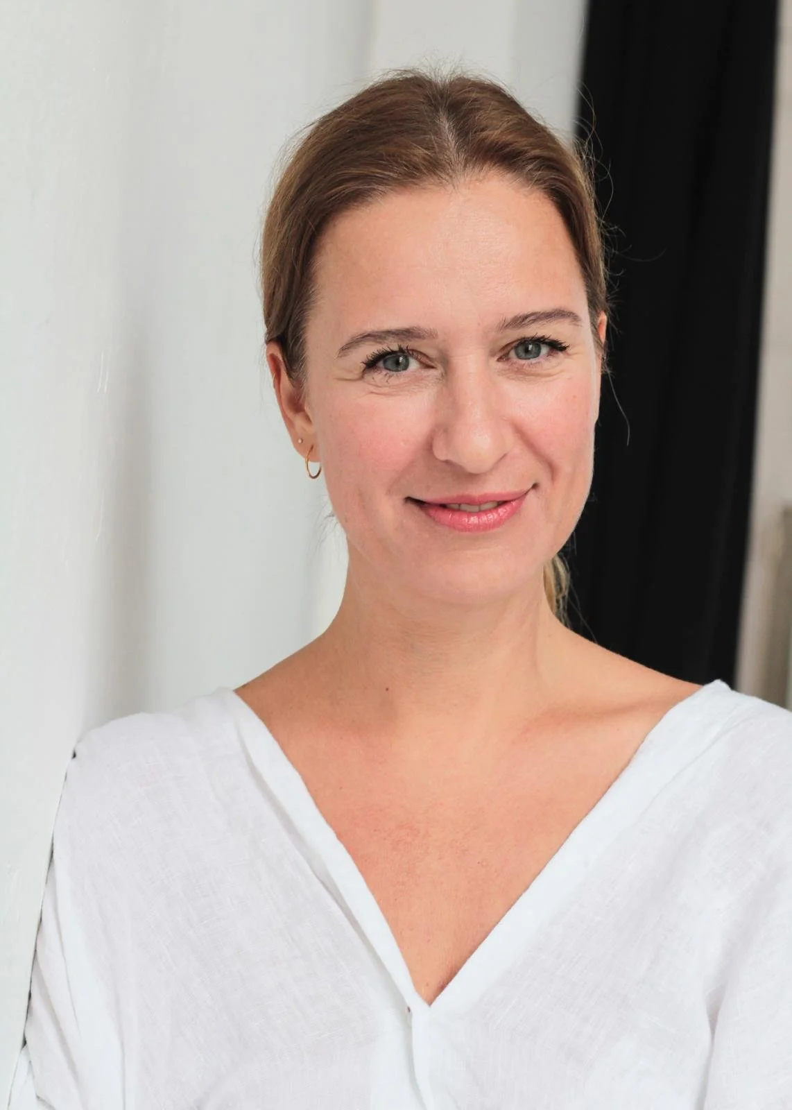 Ein Porträt einer Frau mit blauen Augen, braunem Haar und einem weißen Oberteil, die vor einer weißen Wand steht.