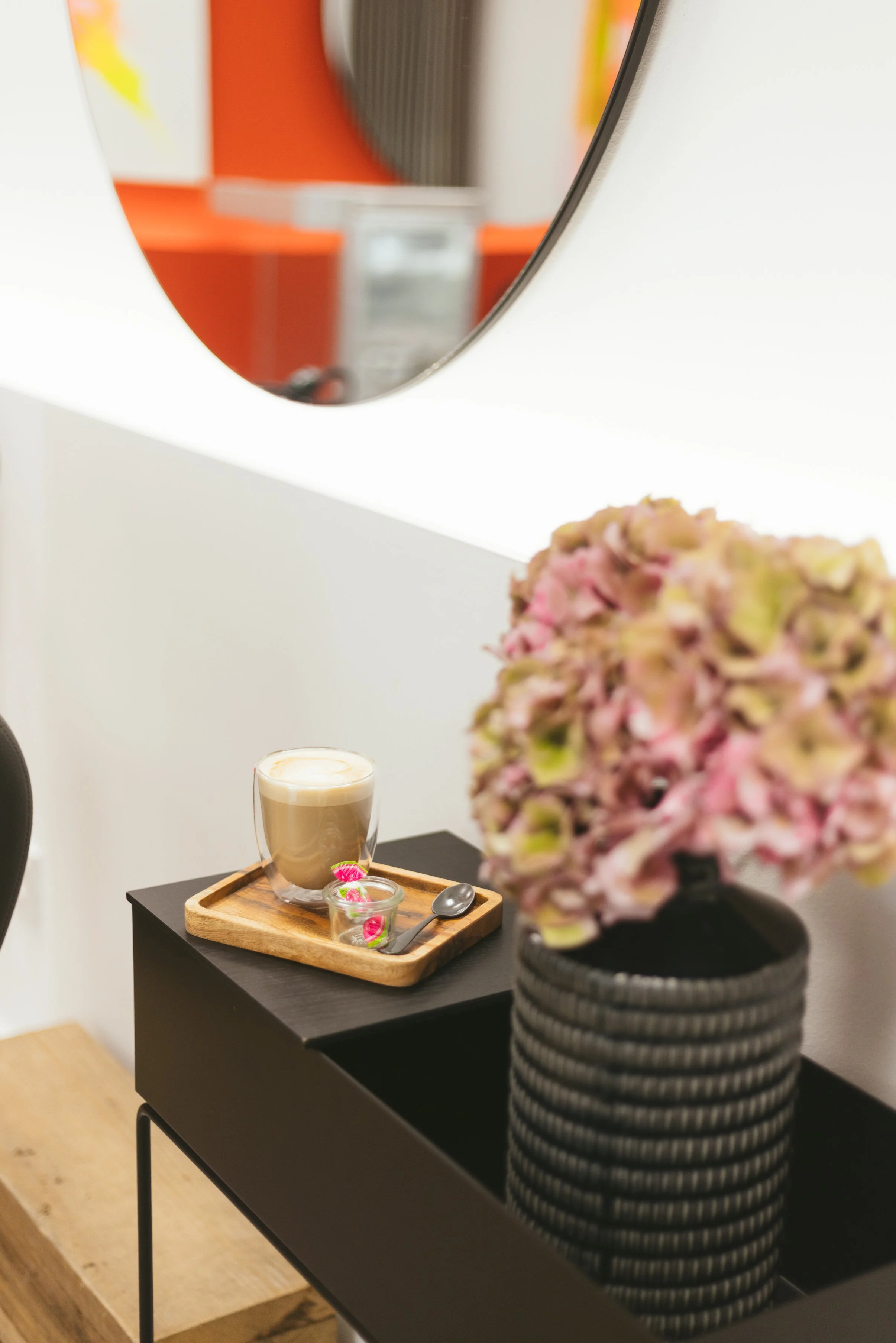 Ein schwarzer Tisch mit einer Holzschale, einem Kaffee in einer Glaskanne, einem Löffel, einer kleinen Schale mit Bonbons und einer Kerze. Im Hintergrund eine Vase mit rosa Blüten. An der Wand ein Spiegel und ein orangefarbener Bereich.