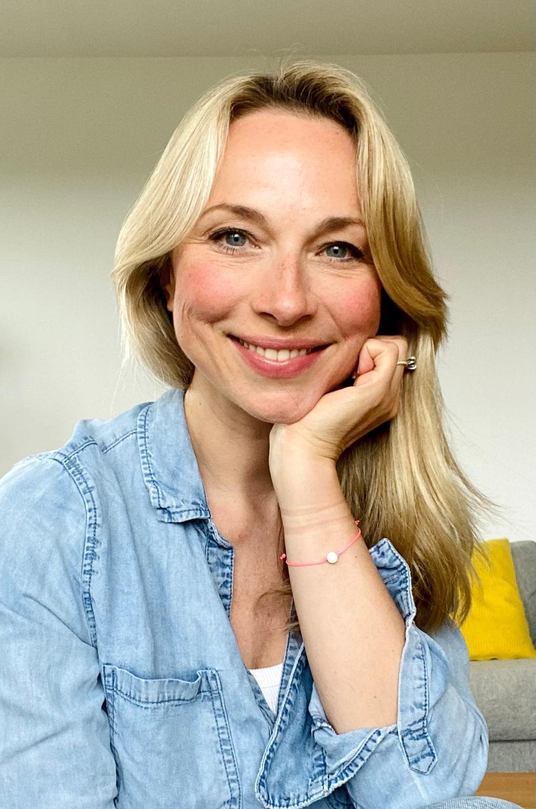 Lächelnde blonde Frau in Jeansjacke, stützt das Kinn an ihrer Hand, sitzt vor einer Couch mit gelbem Kissen, im Innenraum.