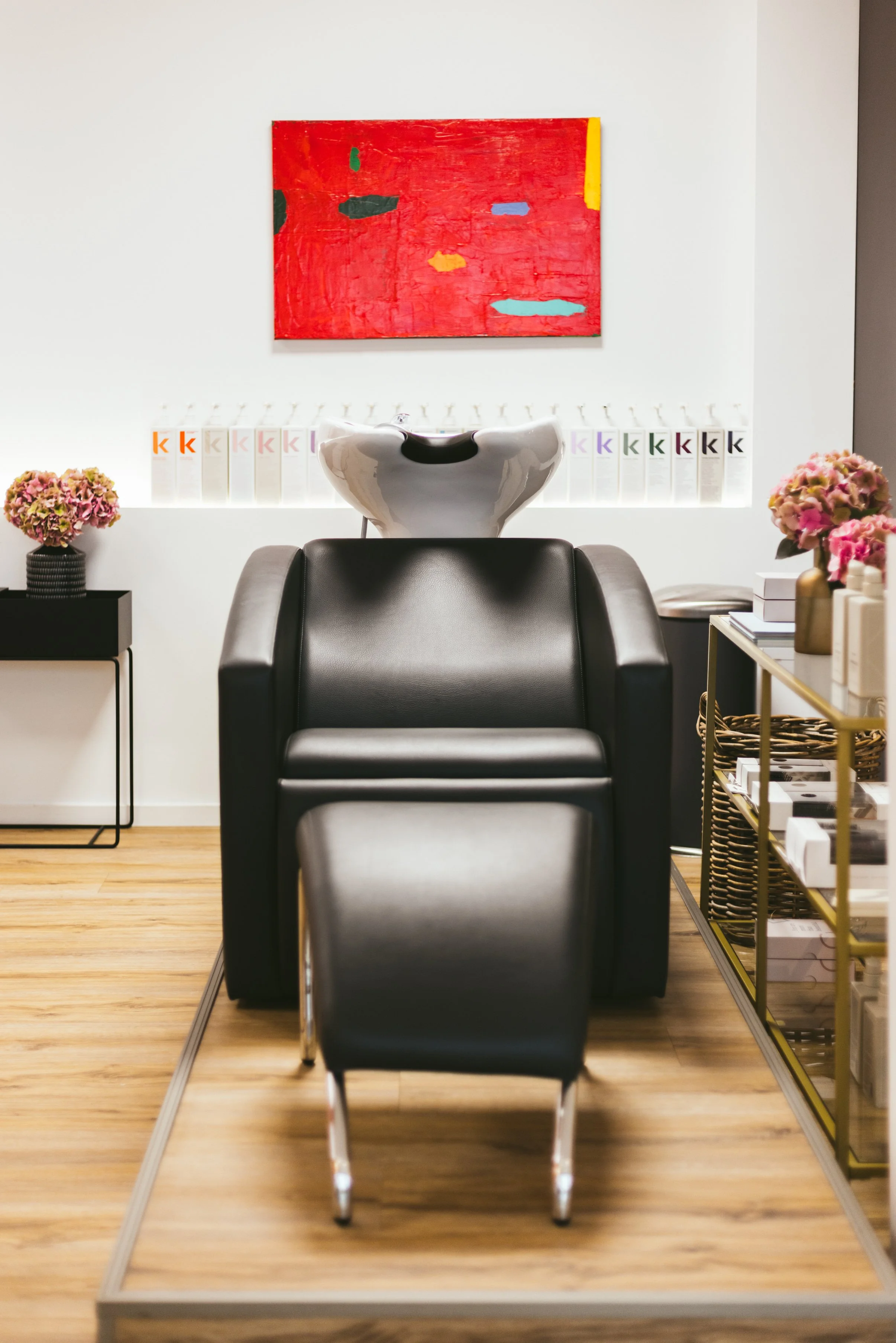 Ein Friseurstuhl vor einem weißen Wand mit einem rot und gelb bemalten Bild. Auf der Ablage befinden sich Haarprodukte und Blumen.