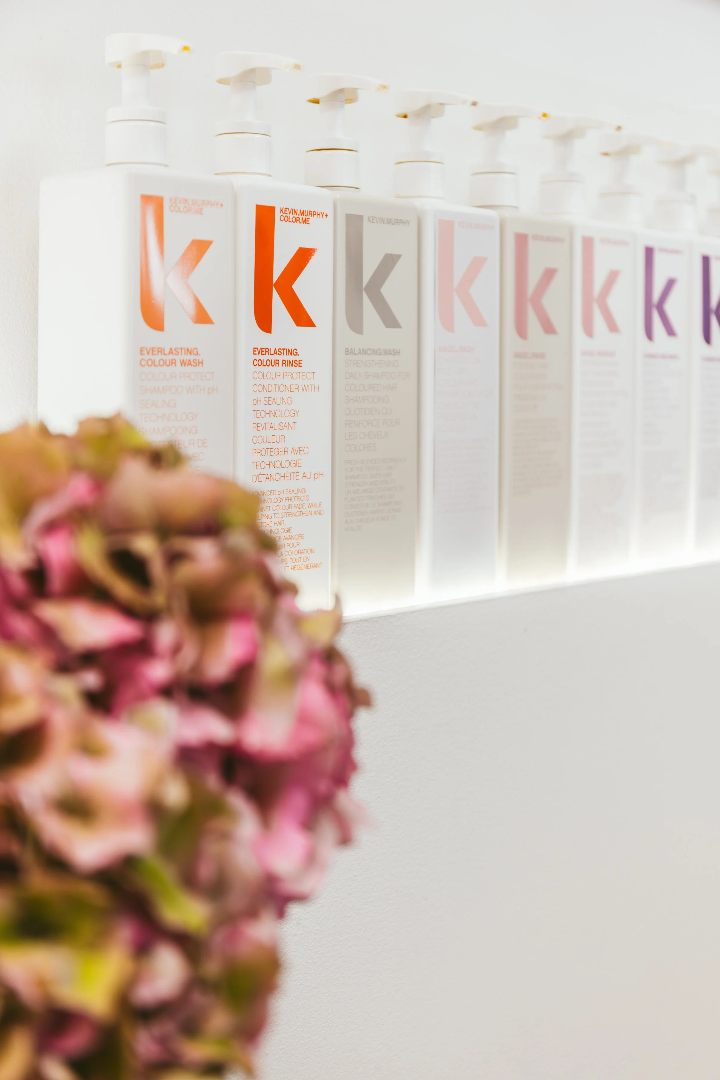 Mehrere weiße Shampoo-Flaschen mit roten, grauen und violetten Labels, die auf einer weißen Wand stehen, vordergründig mit rosa und grünen Blumen im Vordergrund.