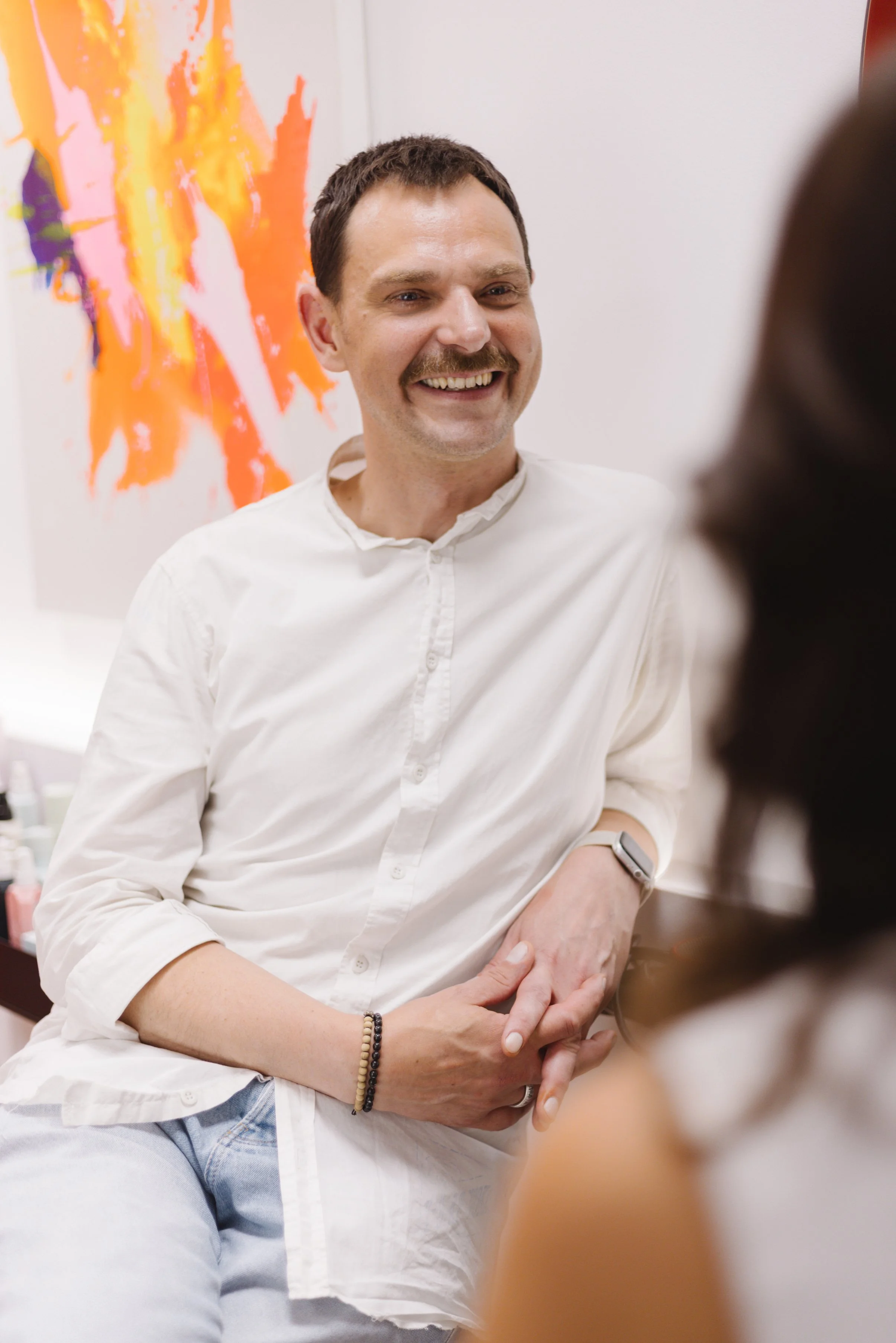 Ein lachender Mann mit dunklem Haar, Bart und Mustache, trägt ein weißes Hemd und sitzt in einem Raum mit einer bunten abstrakten Kunstinstallation im Hintergrund. Er gehört zu einer Unterhaltung mit einer Person im Vordergrund.
