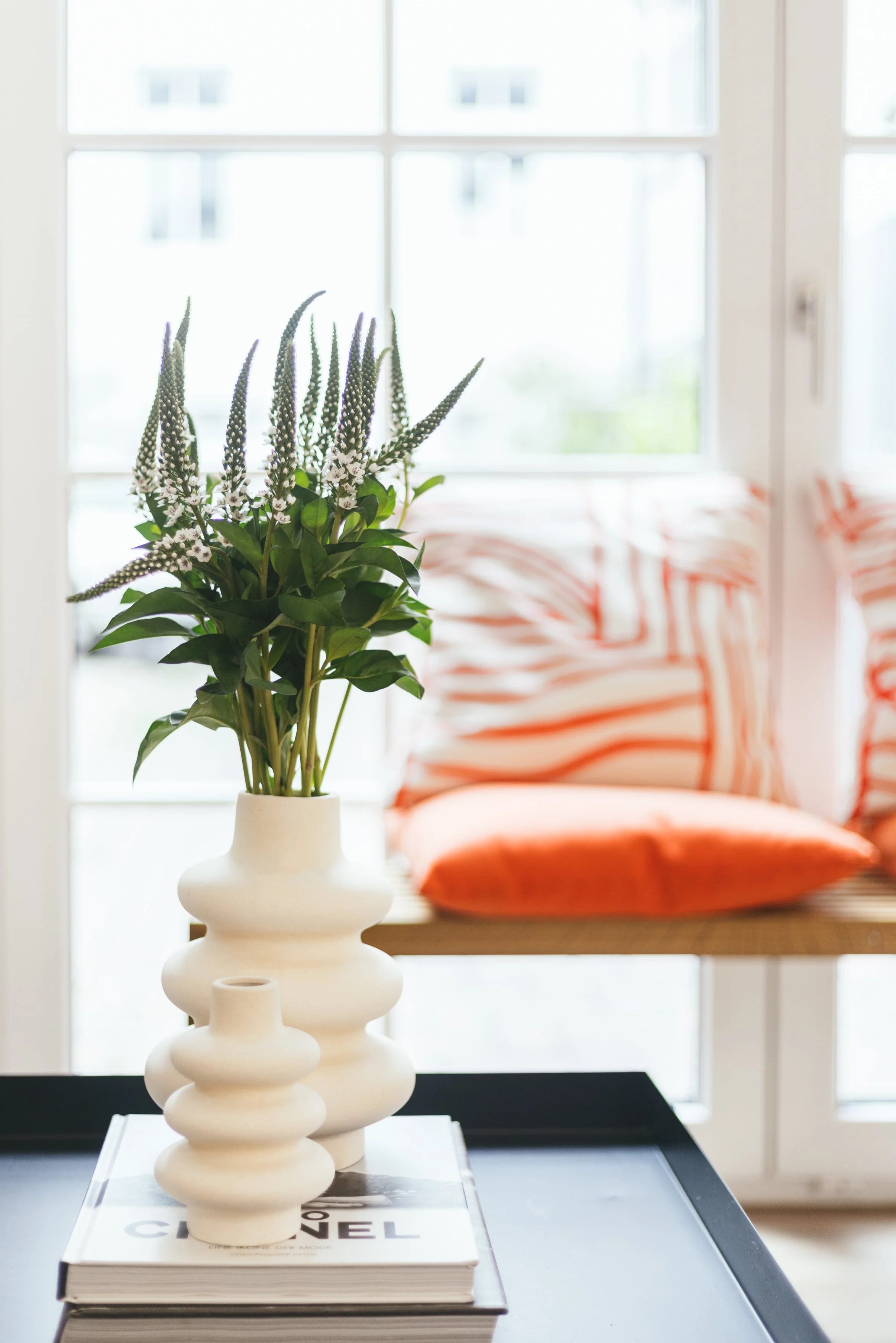 Weiße Vase mit grünen Blättern und weißen und lila Blumen auf einem Buch, im Hintergrund eine Fenster mit Blick nach draußen und orange-weiß gestreifte Kissen auf einem Sofa.