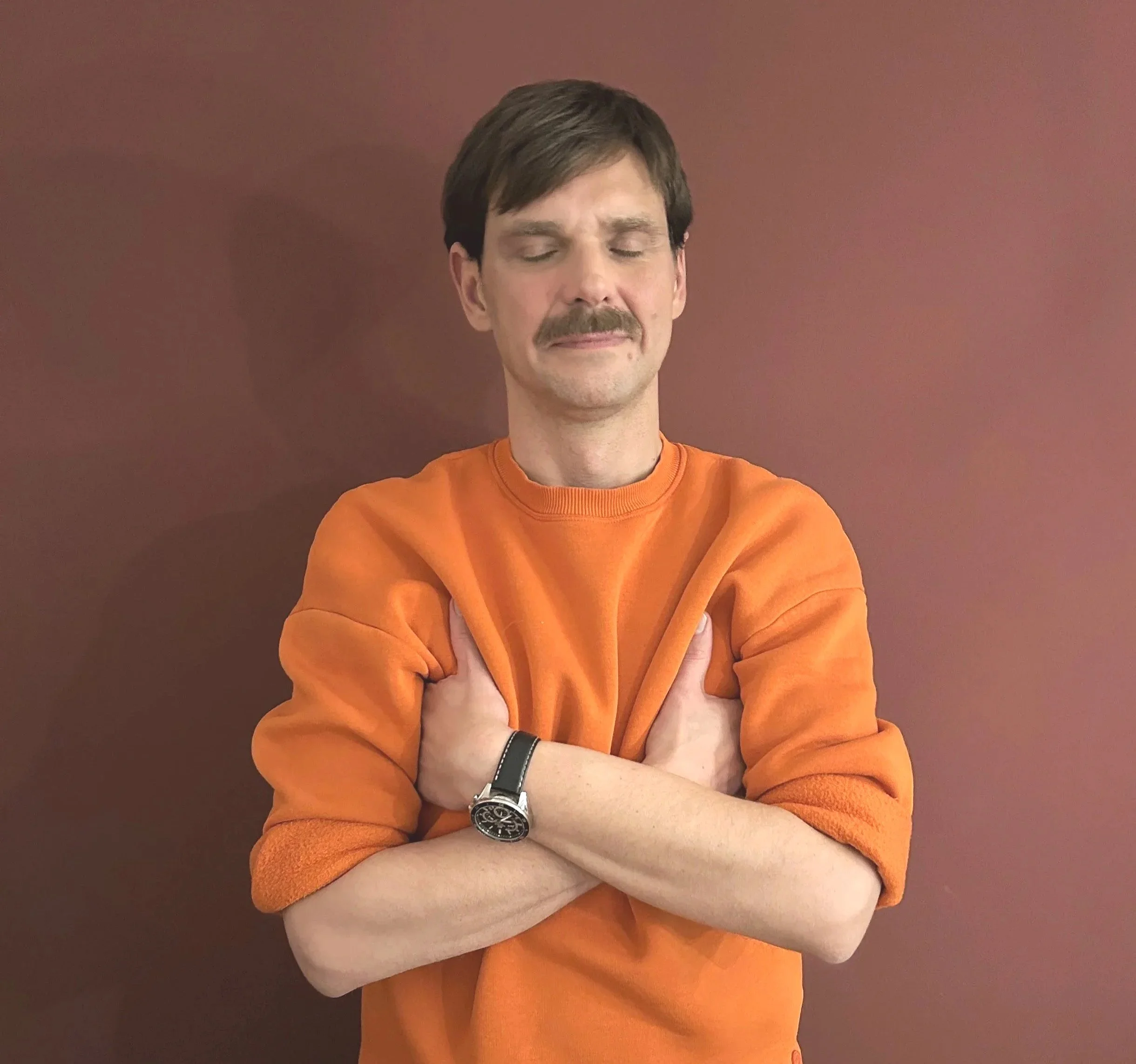 Ein Mann mit dunklem Haar, tBart und geschlossenen Augen, trägt ein orangefarbenes Sweatshirt, steht vor einer braunen Wand, die Arme sind verschränkt. Zeigt einen Jin Shin Jyutsu Griff