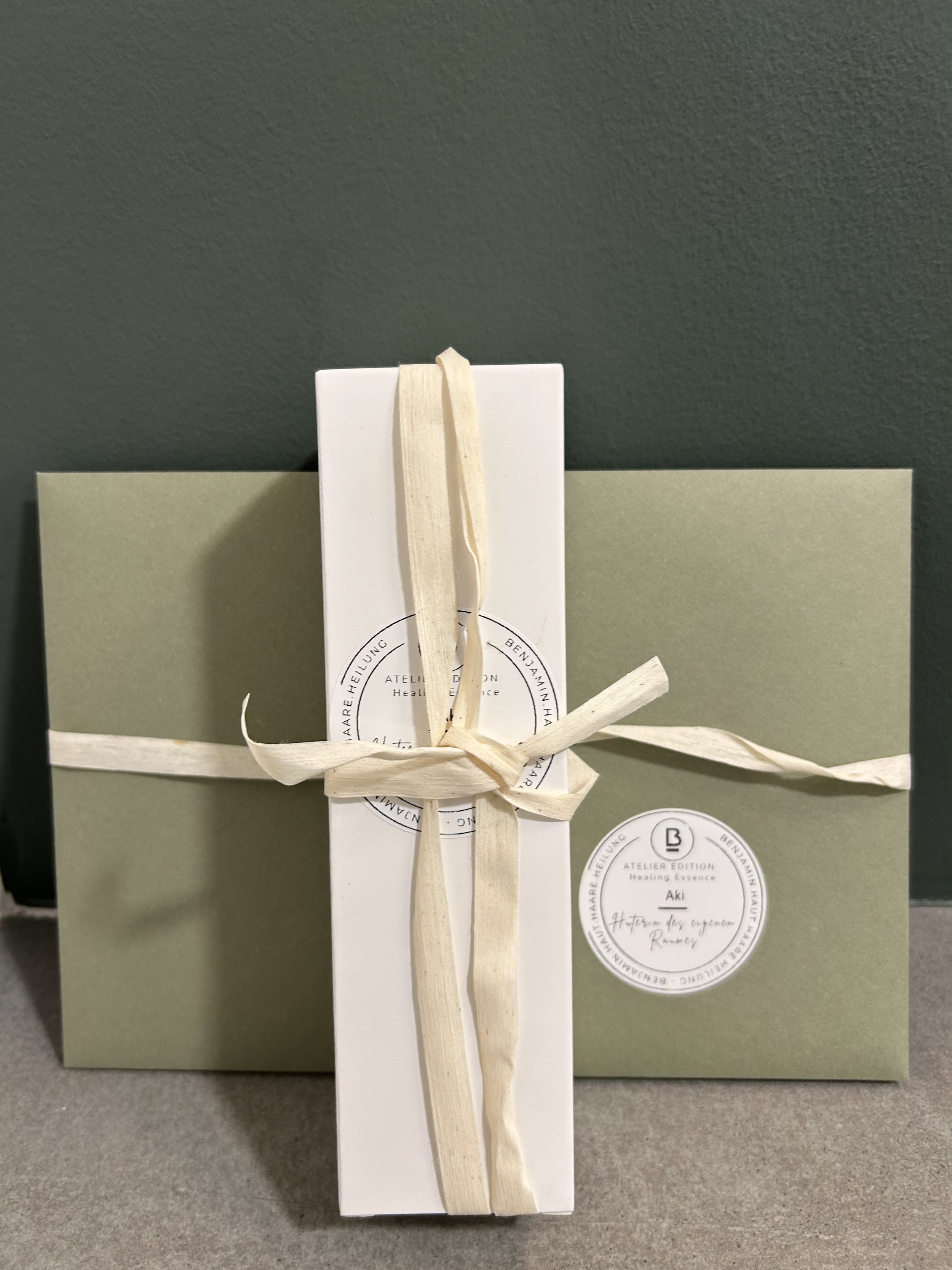 Geschenk mit einer weißen Schleife, in einer weißen Box auf einem grauen Tisch, mit einer grünen Geschenkverpackung im Hintergrund. Es ist eine Heilungsessenz mit japanischer Schrift und einem runden Etikett darunter.