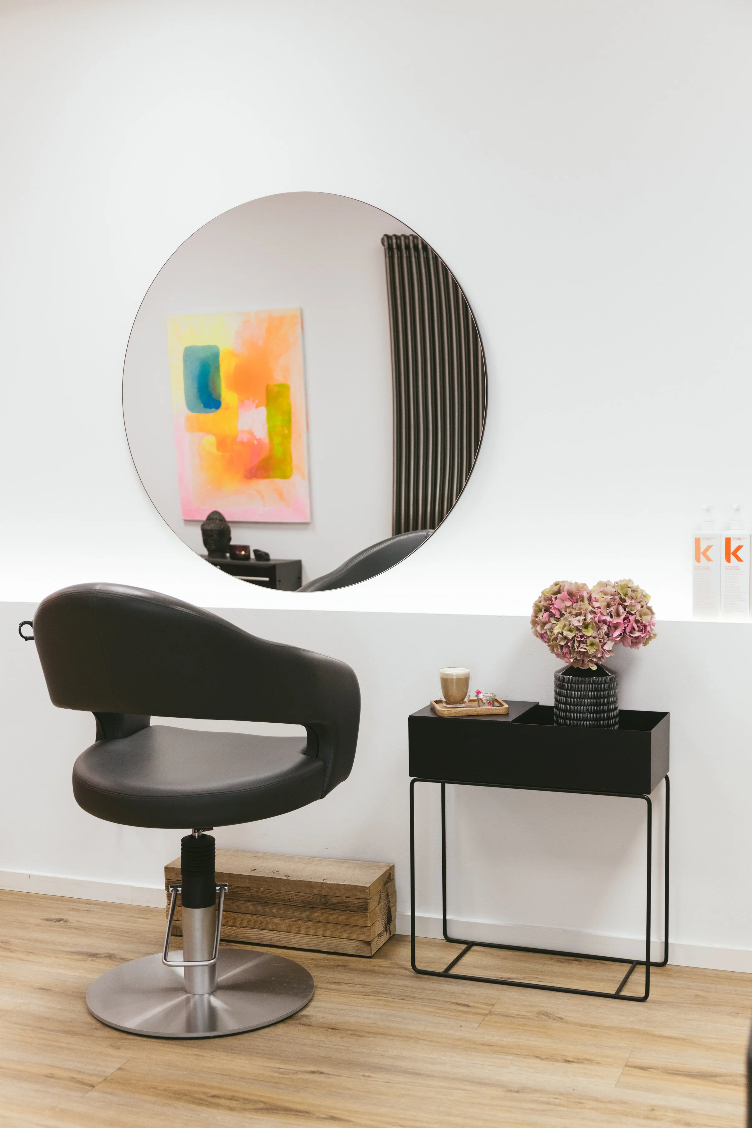 Ein schwarzer Barhocker vor einem weißen Tisch mit Dekoration, darunter eine Vase mit rosa Hortensien, ein Kaminstein und kleine Dekorationsartikel. Im Hintergrund eine grafische Wanddekoration, ein runder Spiegel und ein Heizkörper an der Wand.