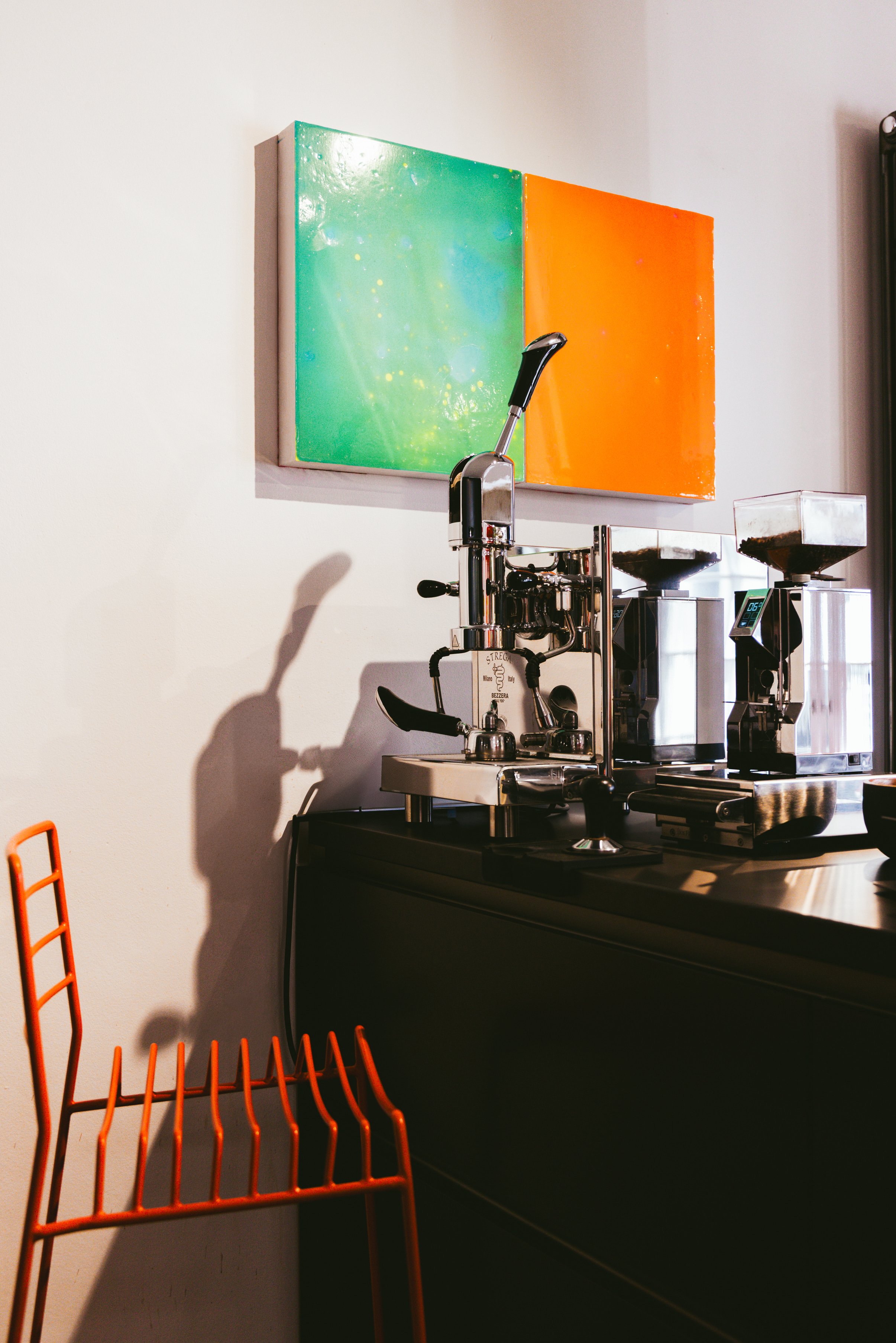 Ein schickes Kaffeezubereitungsgerät auf einer schwarzen Theke, dahinter hängen zwei farbige, quadratische Gemälde an einer weißen Wand, ein orangefarbenes Regal ist im Vordergrund.