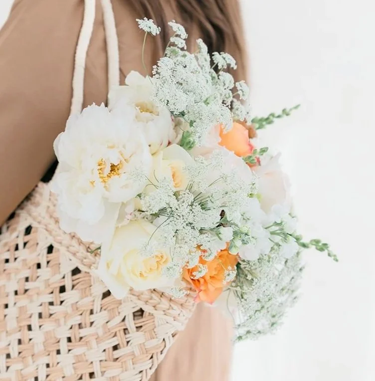 Person holding a bouquet of white and peach flowers, with a woven bag, against a light background.