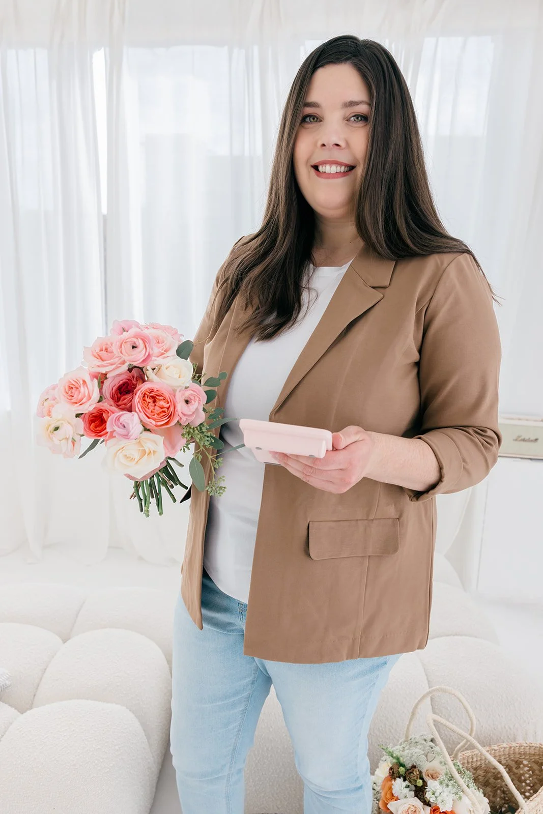 A woman with long brown hair, wearing a beige blazer and light blue jeans, smiling while holding a bouquet of pink, white, and orange roses in one hand and a smartphone in the other, standing in a bright, airy room with white curtains.