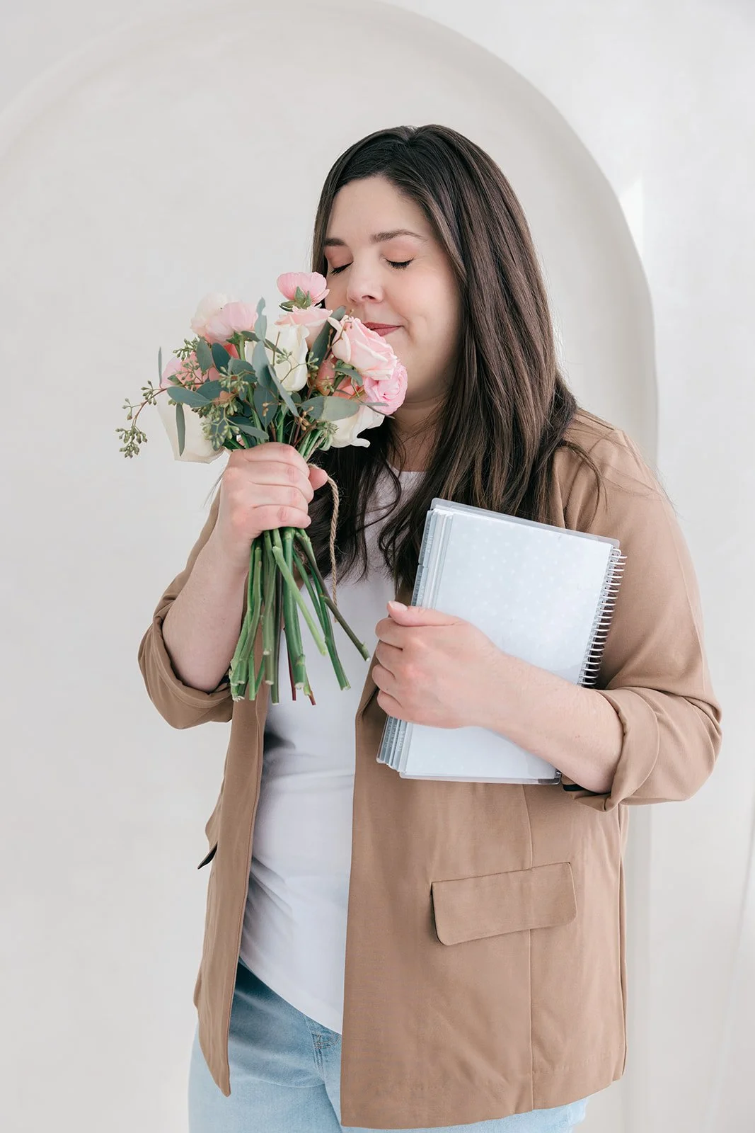 A woman with long dark hair, wearing a tan blazer and white top, is holding a bouquet of pink and white flowers and a notebook, standing in front of a light-colored plain wall.