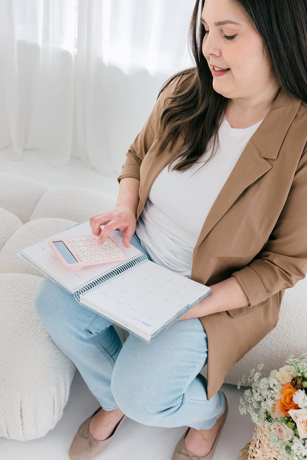 A woman sitting on a white sofa holding a calendar and using a pink calculator, dressed in a tan blazer and light blue jeans, with a basket of flowers nearby.