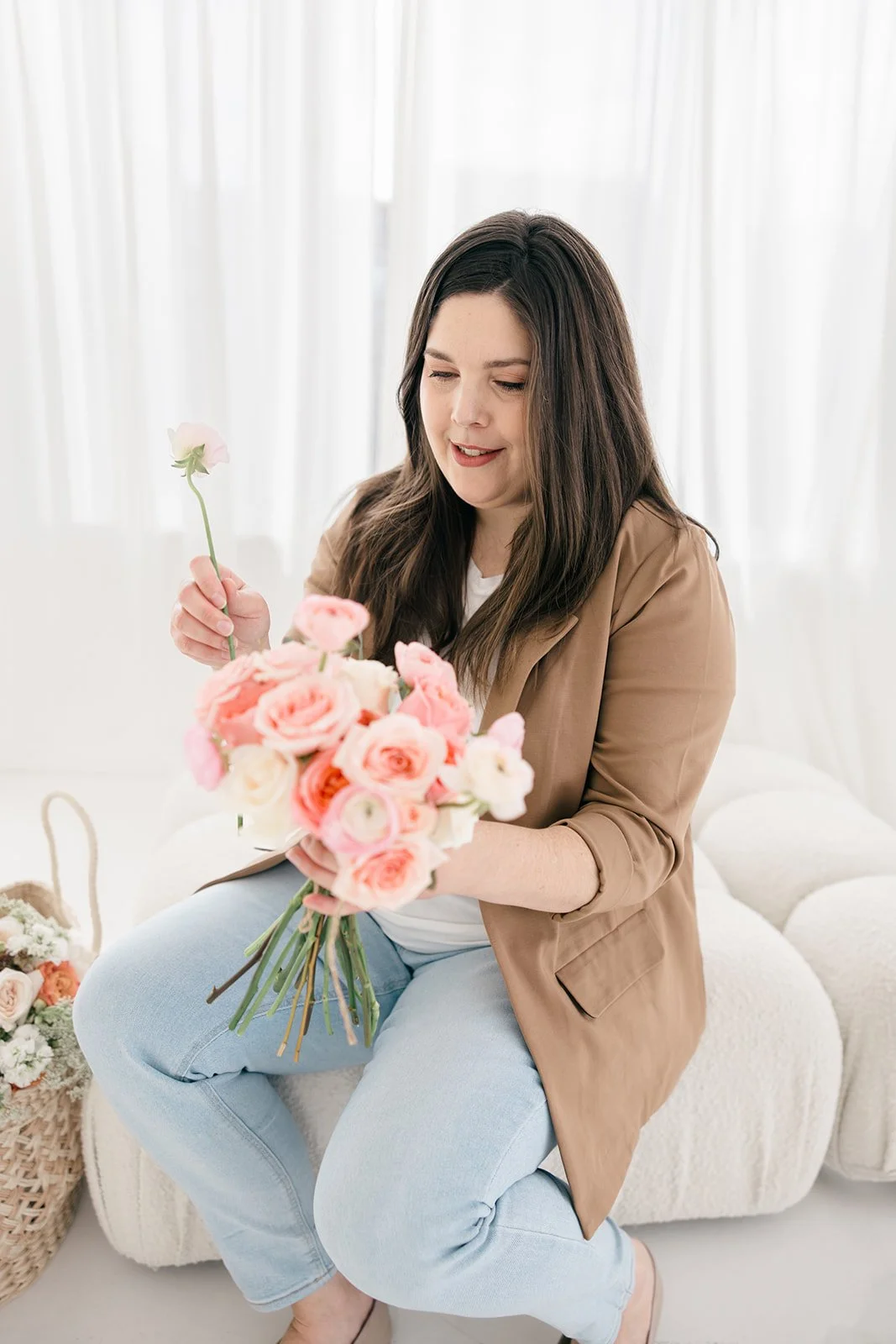 A woman with long dark hair, wearing a light brown blazer and light blue jeans, sitting on a white textured couch, holding and arranging a bouquet of pink and white flowers.