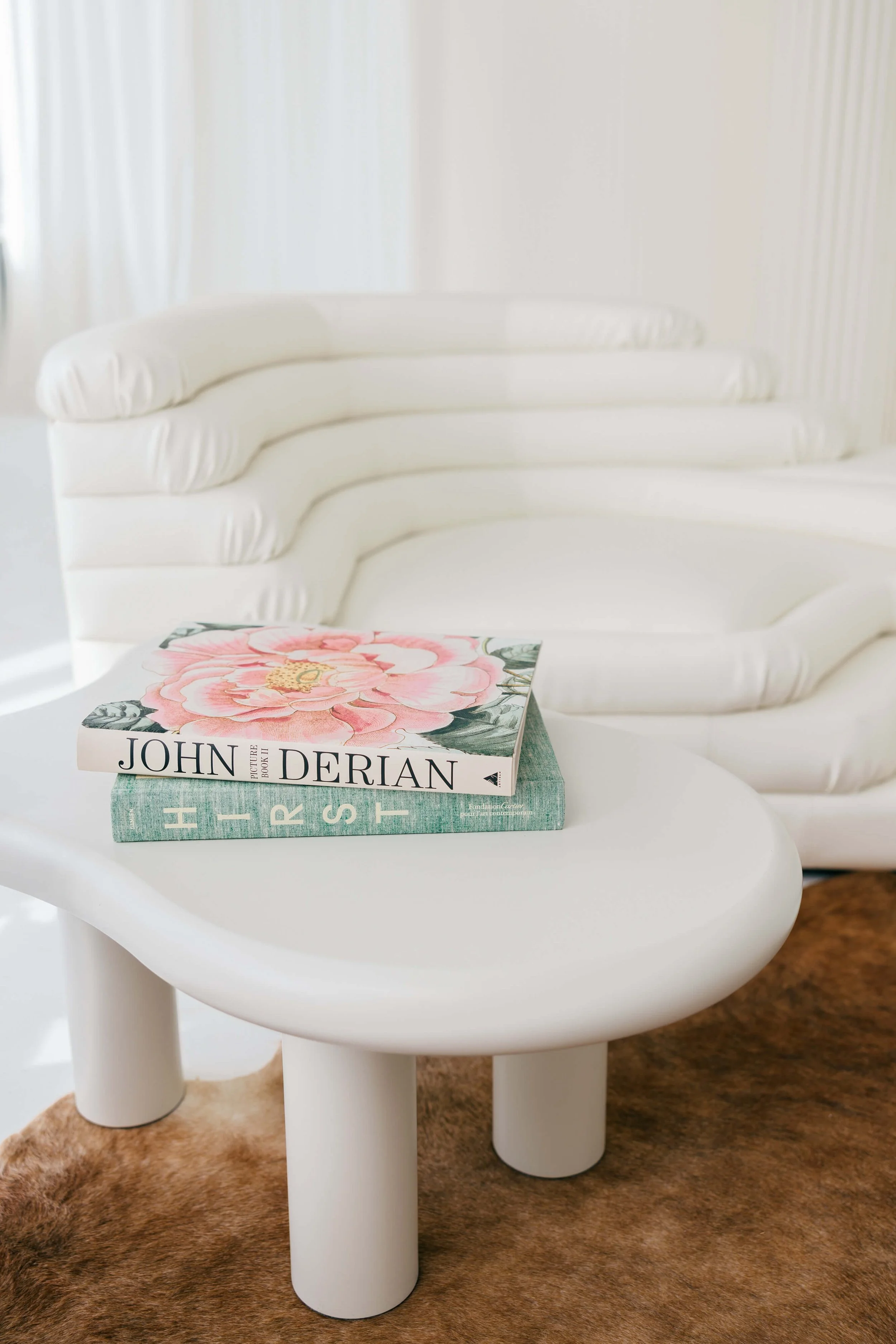 White modern sofa with layered cushions and a low, rounded white coffee table on a brown rug, with two design books stacked on the table, one featuring a pink flower on the cover.