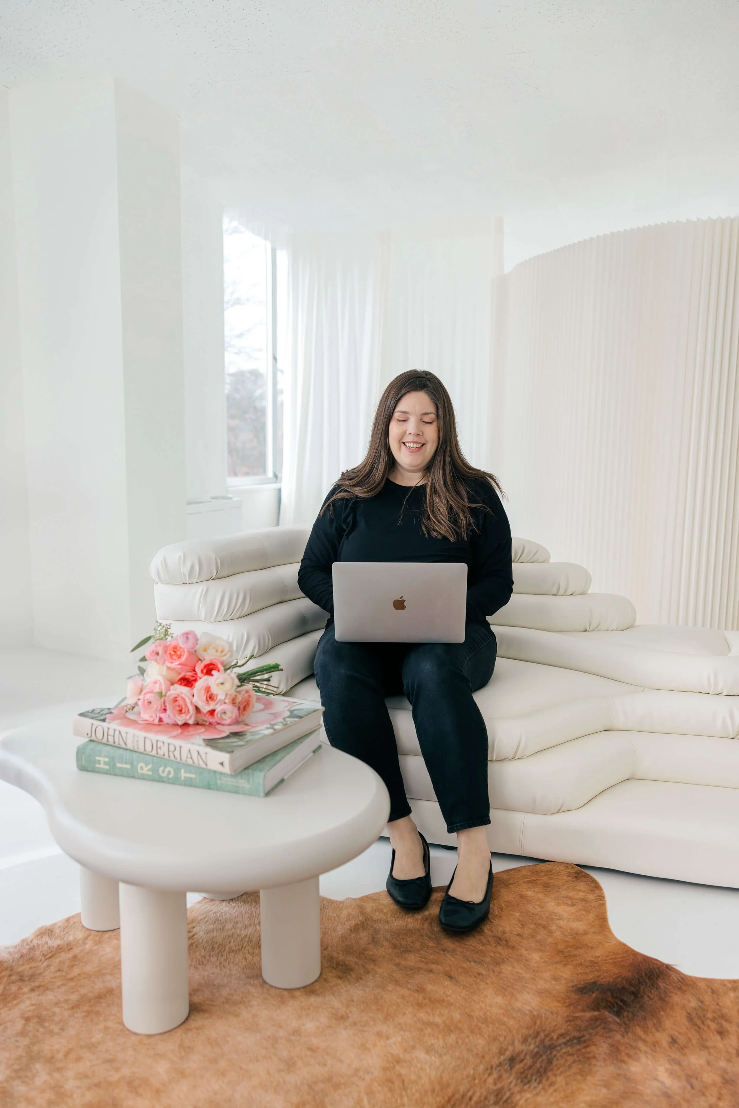 A woman sitting on a white modern sofa with a laptop on her lap, smiling. A coffee table with a bouquet of pink roses and books is in front of her, on top of a cowhide rug. The room has white walls and a large window with white curtains.