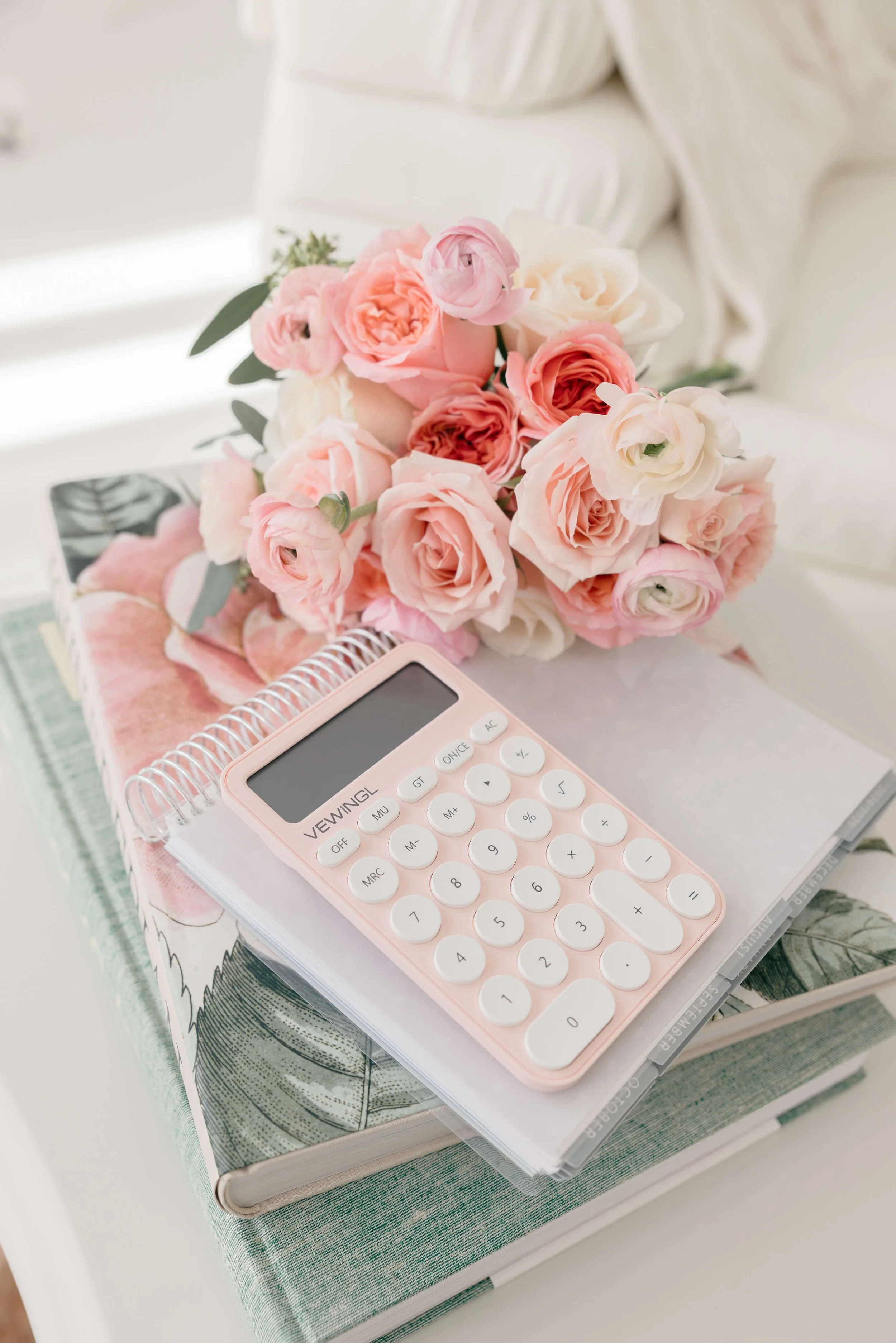 A bouquet of pink and white flowers, an open notebook, a pink calculator, and a book with botanical illustrations on a white surface.