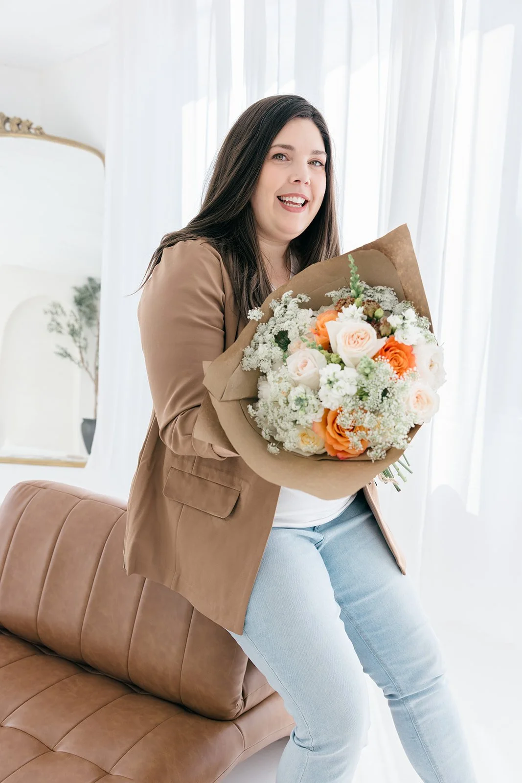 Woman with long dark hair smiling and holding a bouquet of white and orange flowers in a bright room.