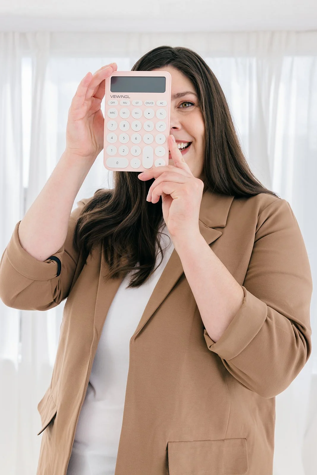 A woman with long dark hair smiling, holding a pink calculator up to her face, with her other hand touching her chin, in a bright room with white curtains.