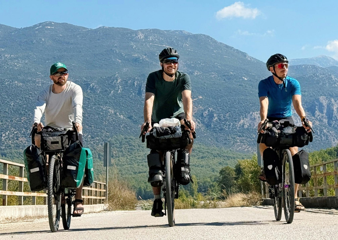 Trois hommes faisant du vélo de route avec sacoches, portant des casques, en montagne par beau temps.