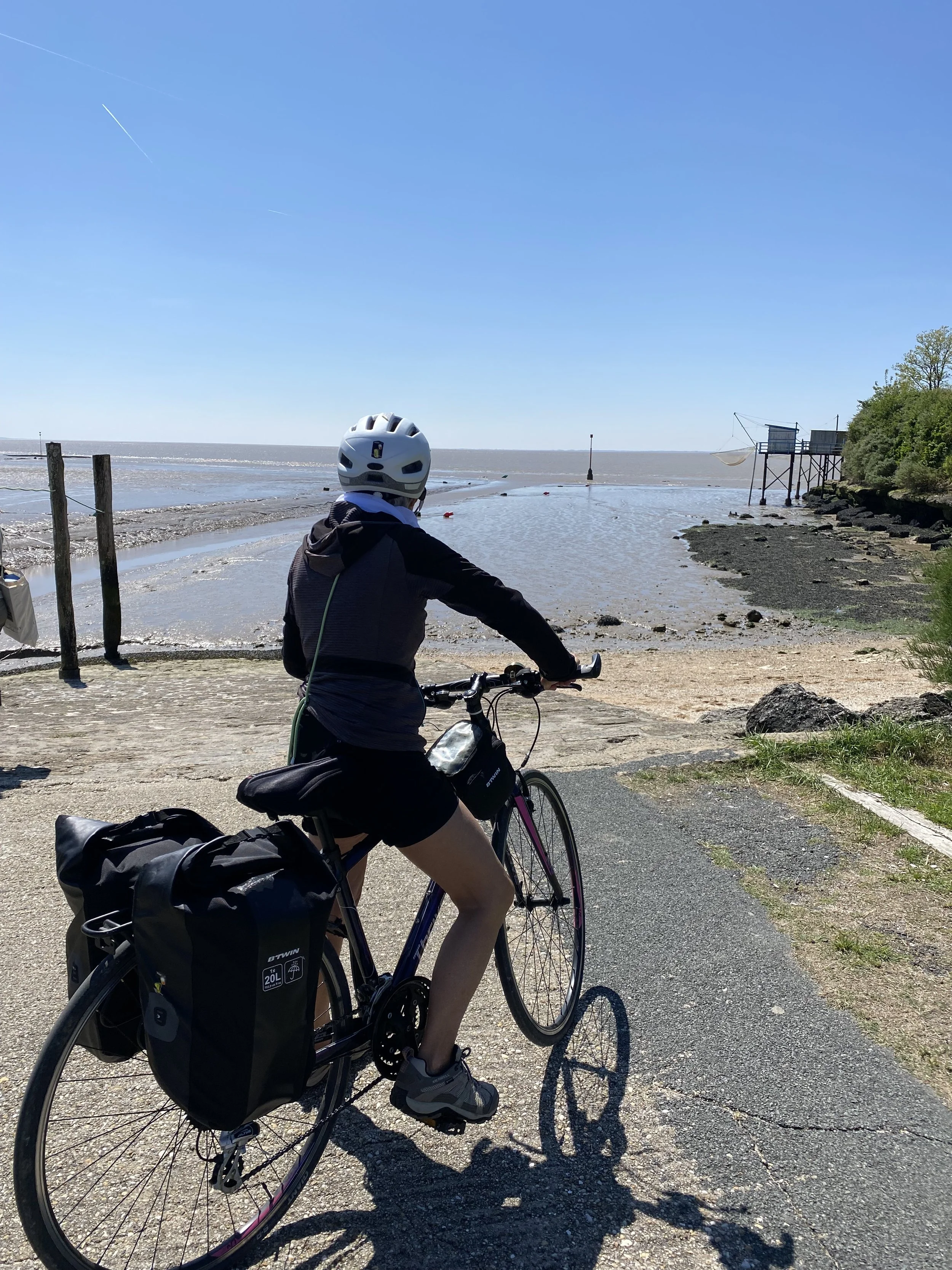 Une personne faisant du vélo le long d'une côte ou d'une plage sous un ciel dégagé, portant un casque blanc, une veste noire, et avec un sac attaché à son vélo.