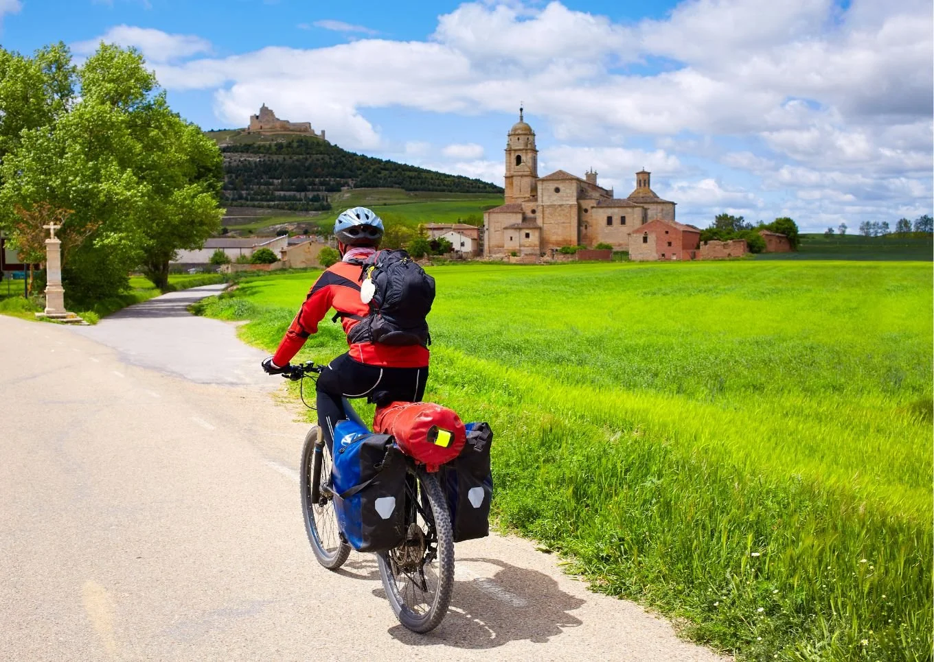 Cycliste avec sacoches, portant un casque, roulant sur un chemin rural avec une église en pierre et un château à l'arrière-plan