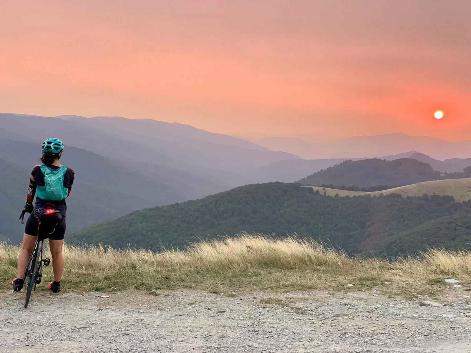 Une personne en vélo, portant un casque bleu, se tient à l'arrêt, regardant un coucher de soleil sur des montagnes verdoyantes, avec un ciel rose et une lumière orange, sur la véloroute Scandibérique.
