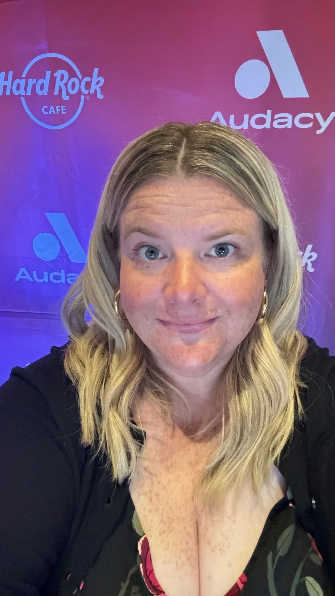 Selfie of a woman with blonde hair and blue eyes, smiling, with a purple background displaying the Hard Rock Cafe and Audacy logos.