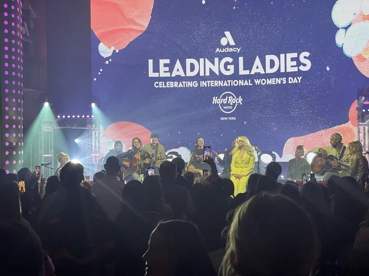 Women musicians performing on stage at a celebration of International Women's Day, with a large screen displaying "Leading Ladies" and "Celebrating International Women's Day," at the Hard Rock Hotel in New York.
