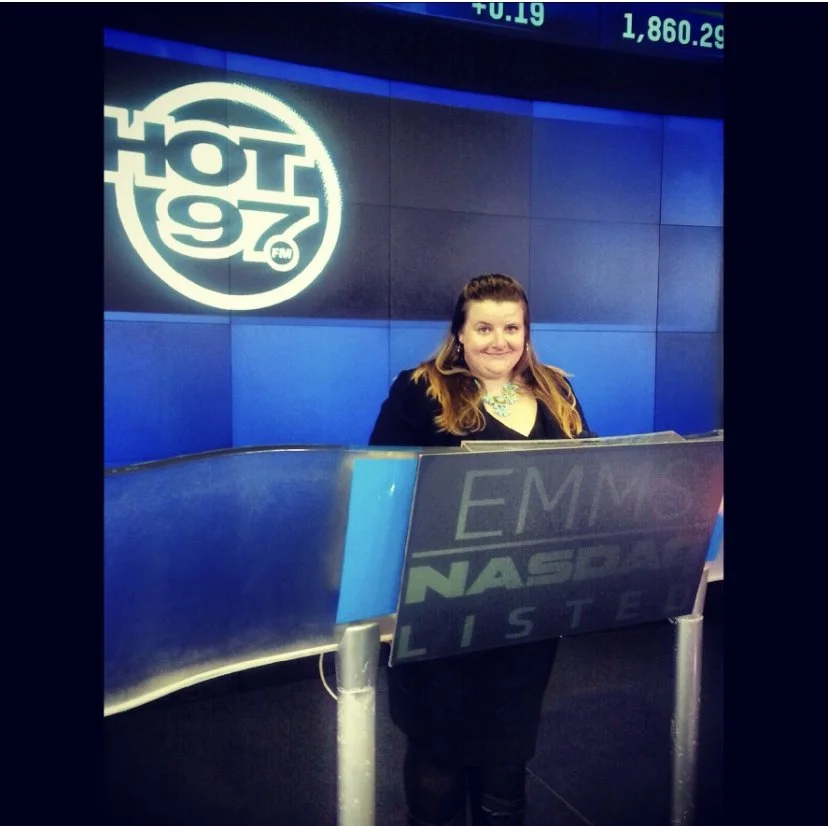 A woman stands behind a reception desk at iHeartRadio with the 97. FM logo on the wall behind her, inside a studio with a digital market display.