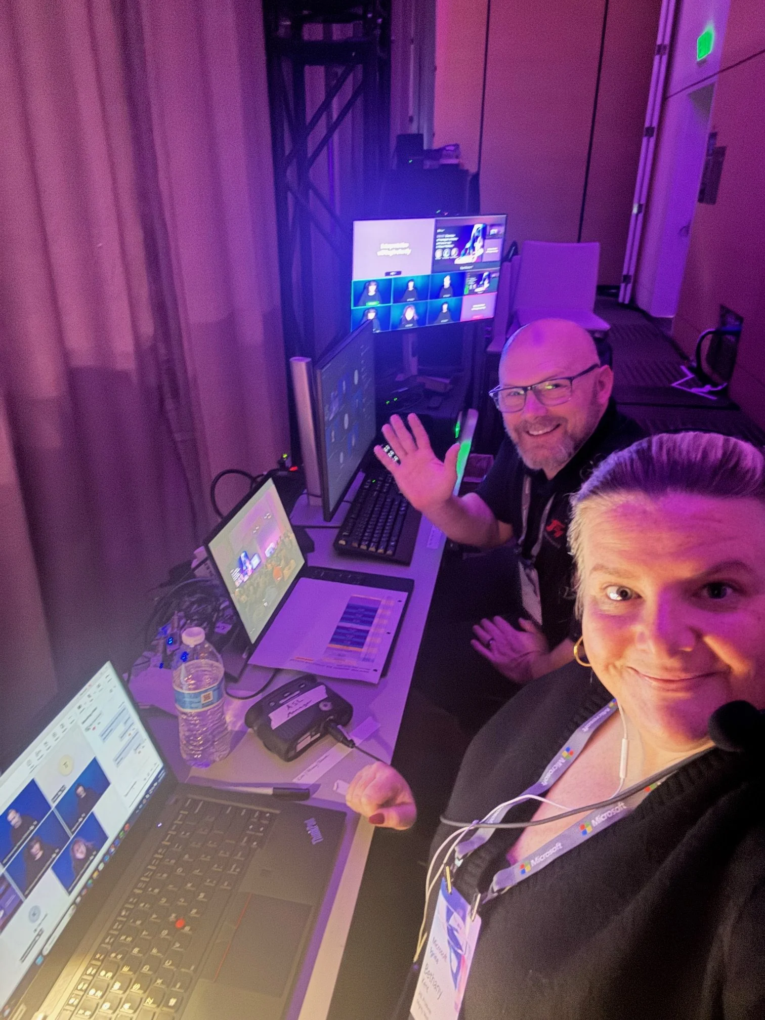 Two people at a technology setup with multiple monitors, laptops, and electronic devices in a room with purple lighting, possibly at a conference or event.