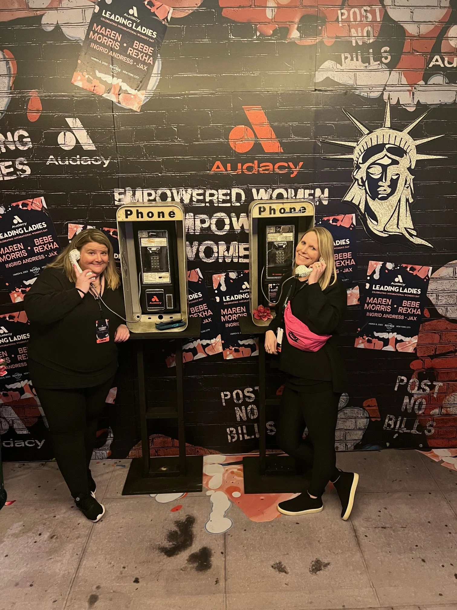 Two women stand at payphones, smiling and talking on the phones. The background features a brick wall decorated with posters and artwork promoting Women's Day, with slogans like "Powered Women" and the logo "Audacy". The women are wearing black outfi