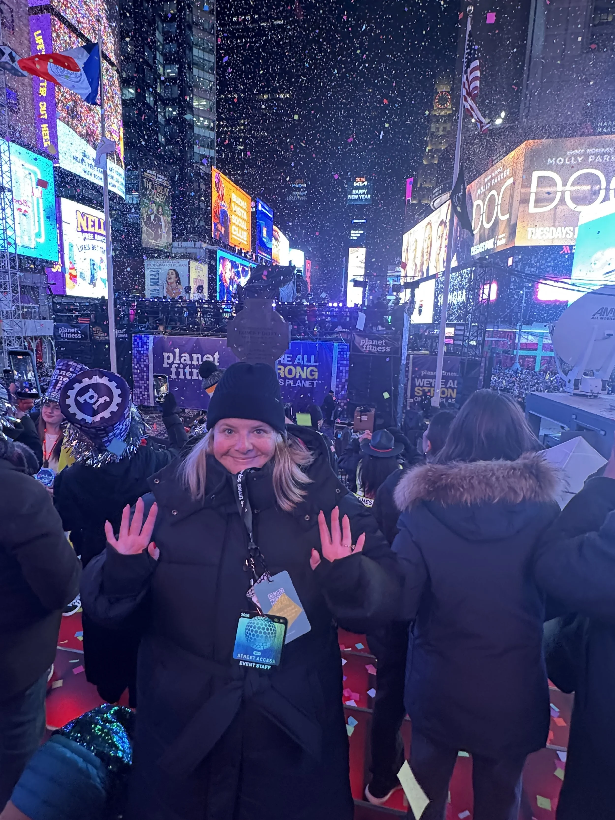 A woman smiling and holding up her hands in Times Square, New York City, during a celebration with confetti falling around her, bright neon signs, billboards, and crowds in the background.